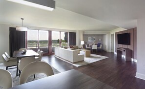 A modern hotel room with a dining area, a living room with a sofa and chairs, a flat-screen TV mounted on the wall, and large windows offering a view of the outdoors.