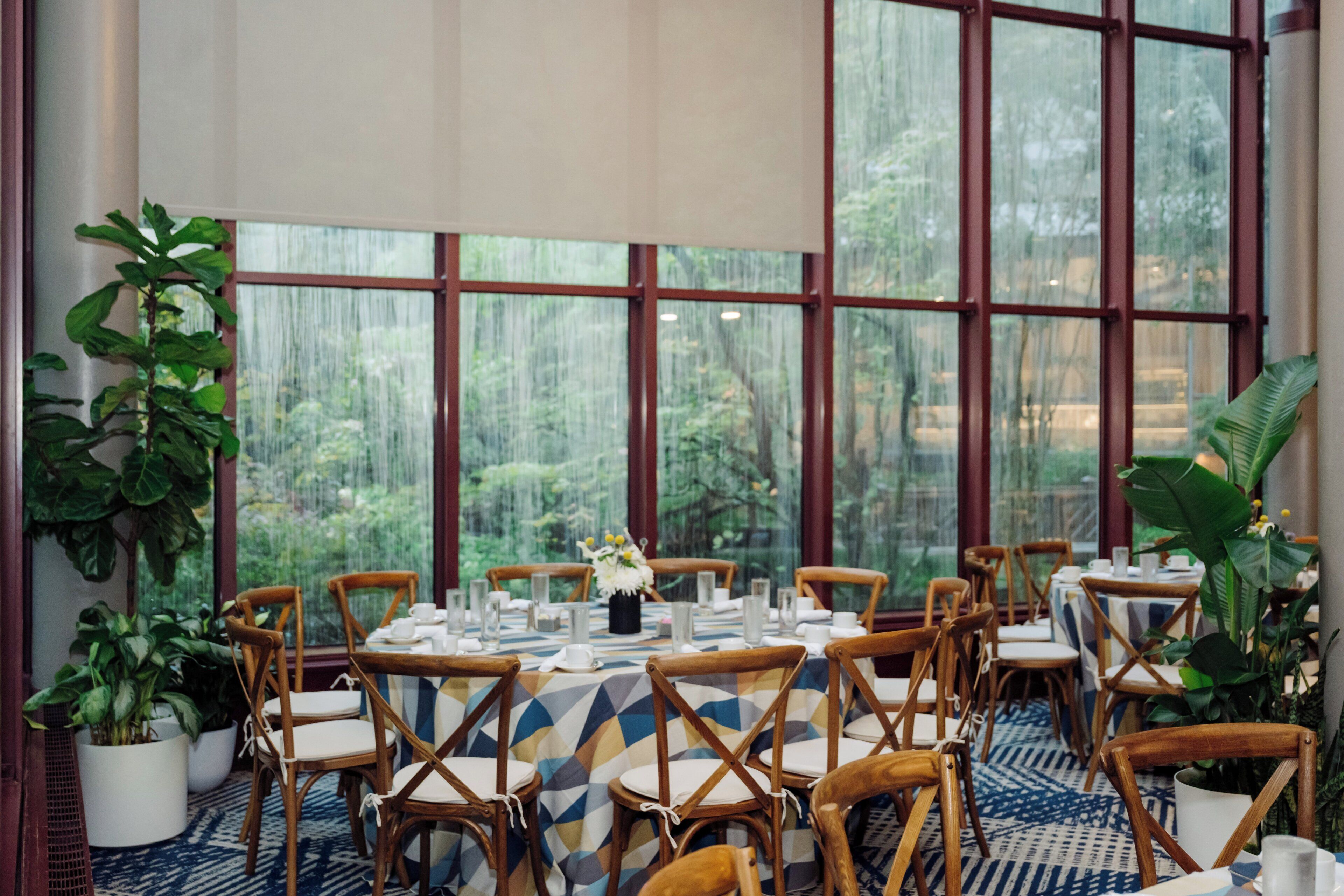A dining area with tables set for six, surrounded by wooden chairs, large windows with a view of greenery, and potted plants.