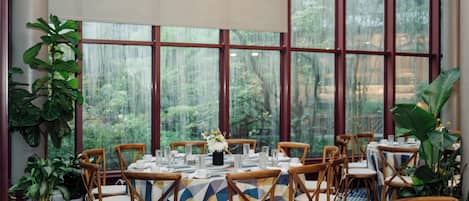 A dining area with tables set for six, surrounded by wooden chairs, large windows with a view of greenery, and potted plants.