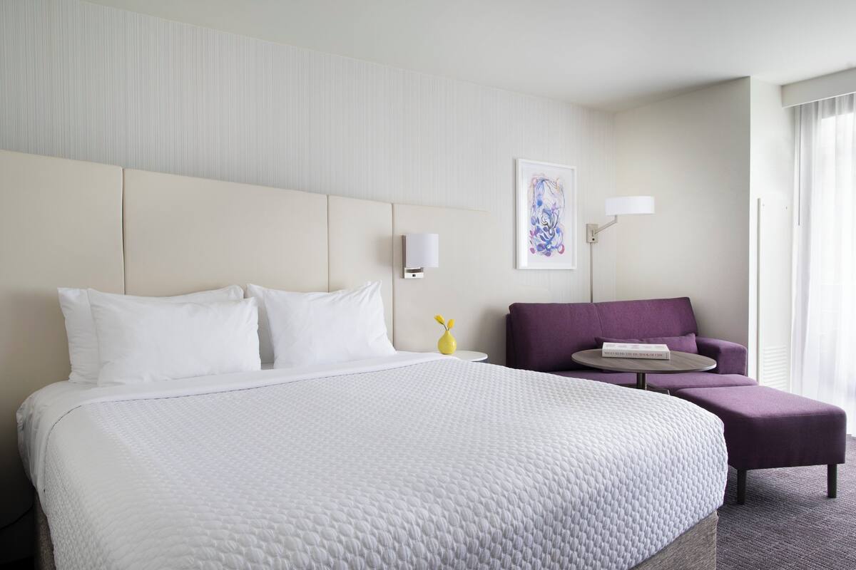 A hotel room with a large bed, a small sofa, a round table, and a wall-mounted lamp.