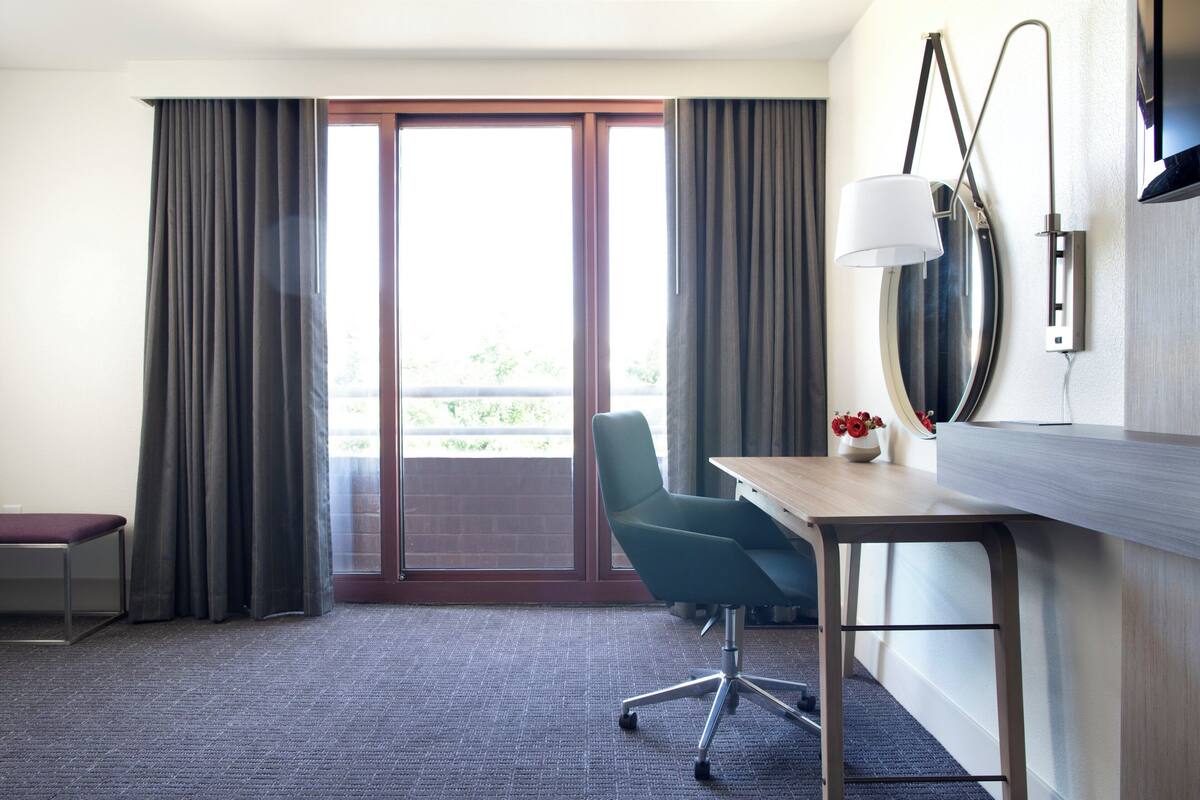 Habitación de hotel con escritorio, silla y un amplio ventanal con cortinas.