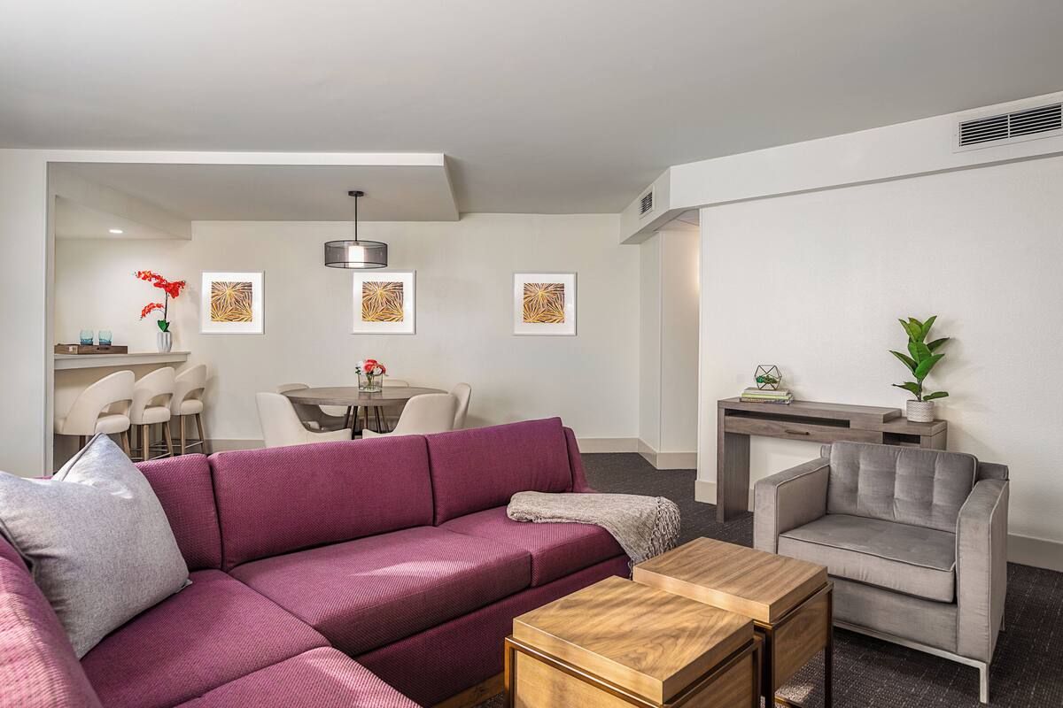 A modern living room with a purple sofa, a grey armchair, and a wooden coffee table. There is a dining area with a table and chairs in the background.