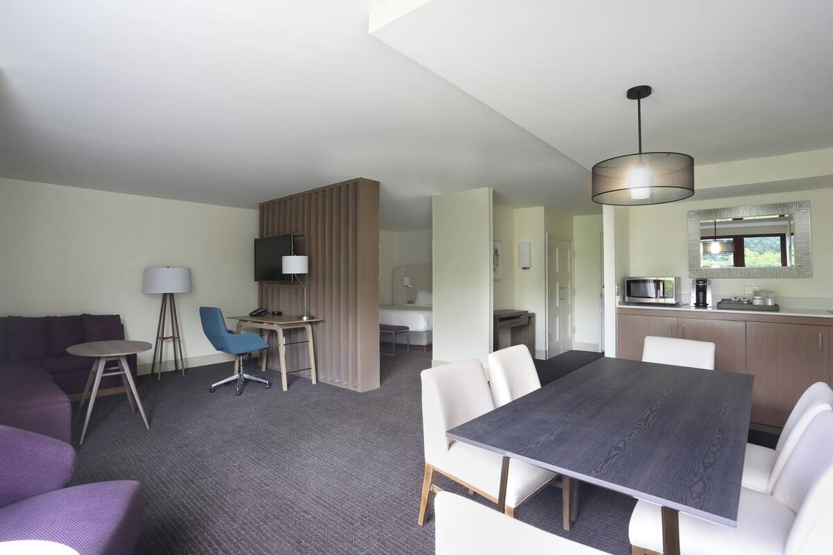 A modern hotel room with a dining area, a sofa, a small desk, and a kitchenette.