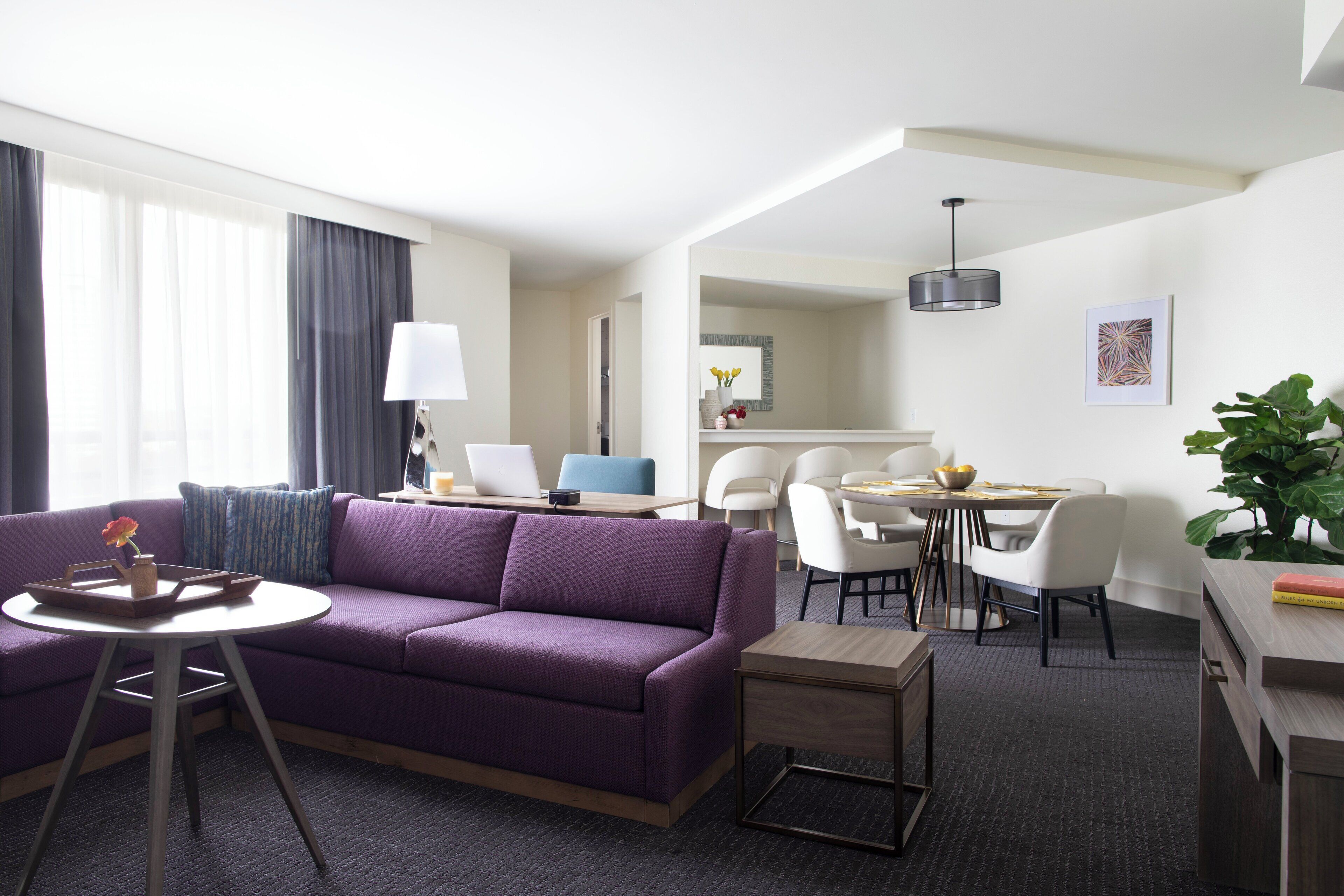 A modern hotel room with a purple sofa, a round dining table, white chairs, a small side table, and a large window with curtains.