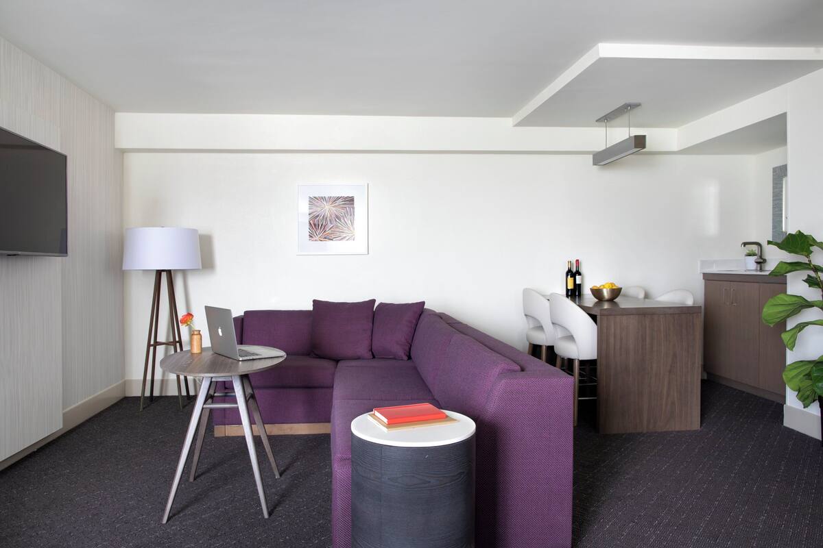 A modern living room with a purple sofa, a round coffee table, a flat-screen TV, and a small dining area with a white chair and a wooden sideboard.