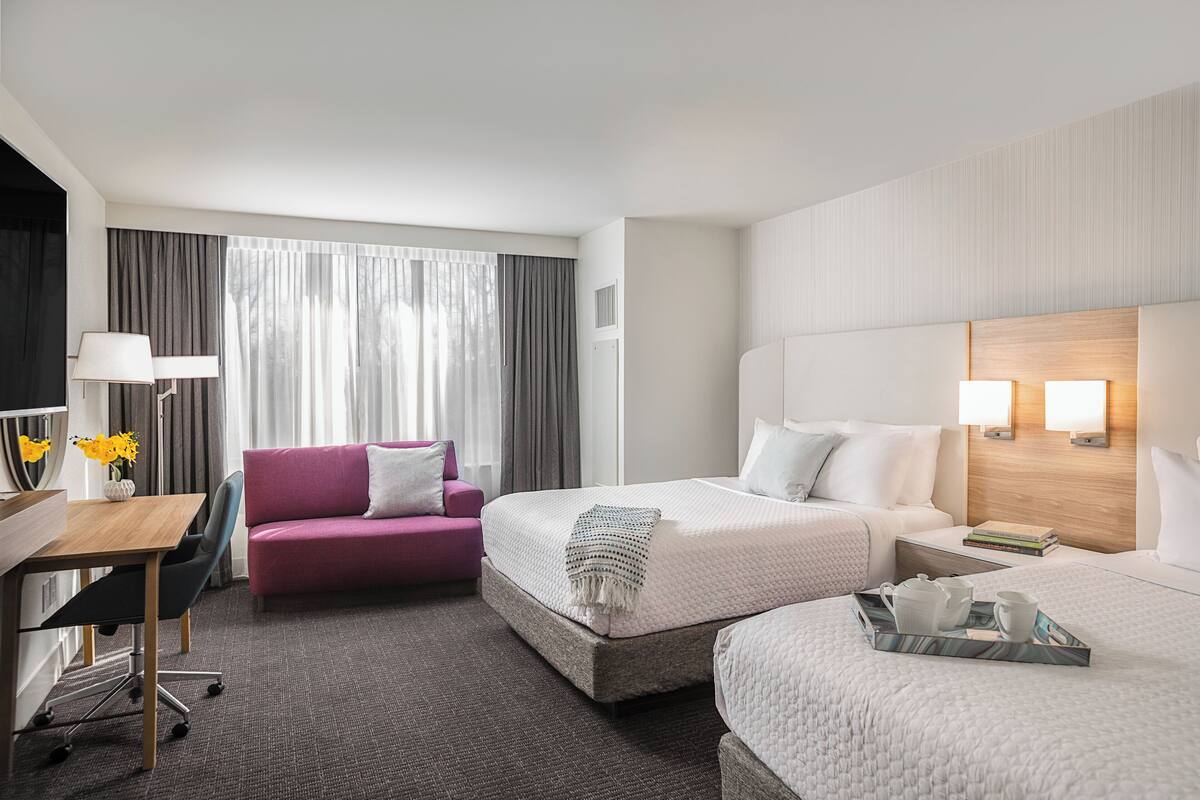 A hotel room with a bed, a desk, a chair, a sofa, and a bedside table with books and a tray.