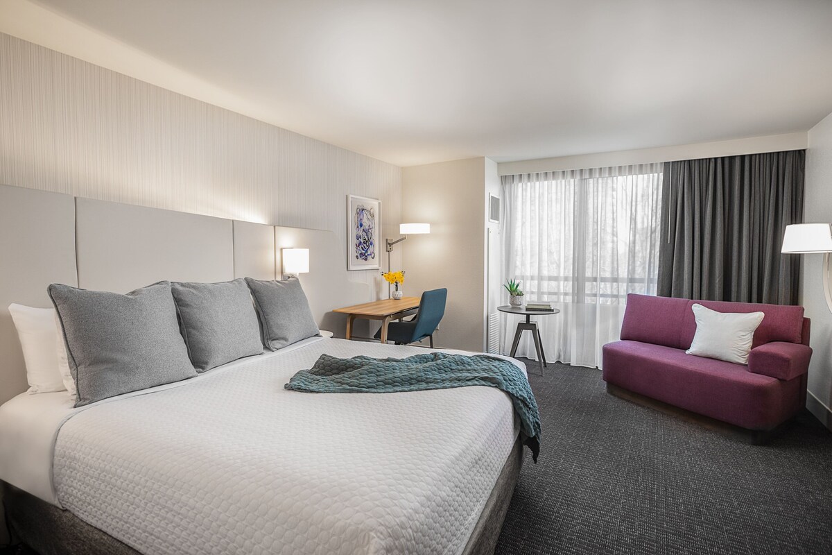 A hotel room with a large bed, a desk, a chair, a sofa, and a window with curtains.
