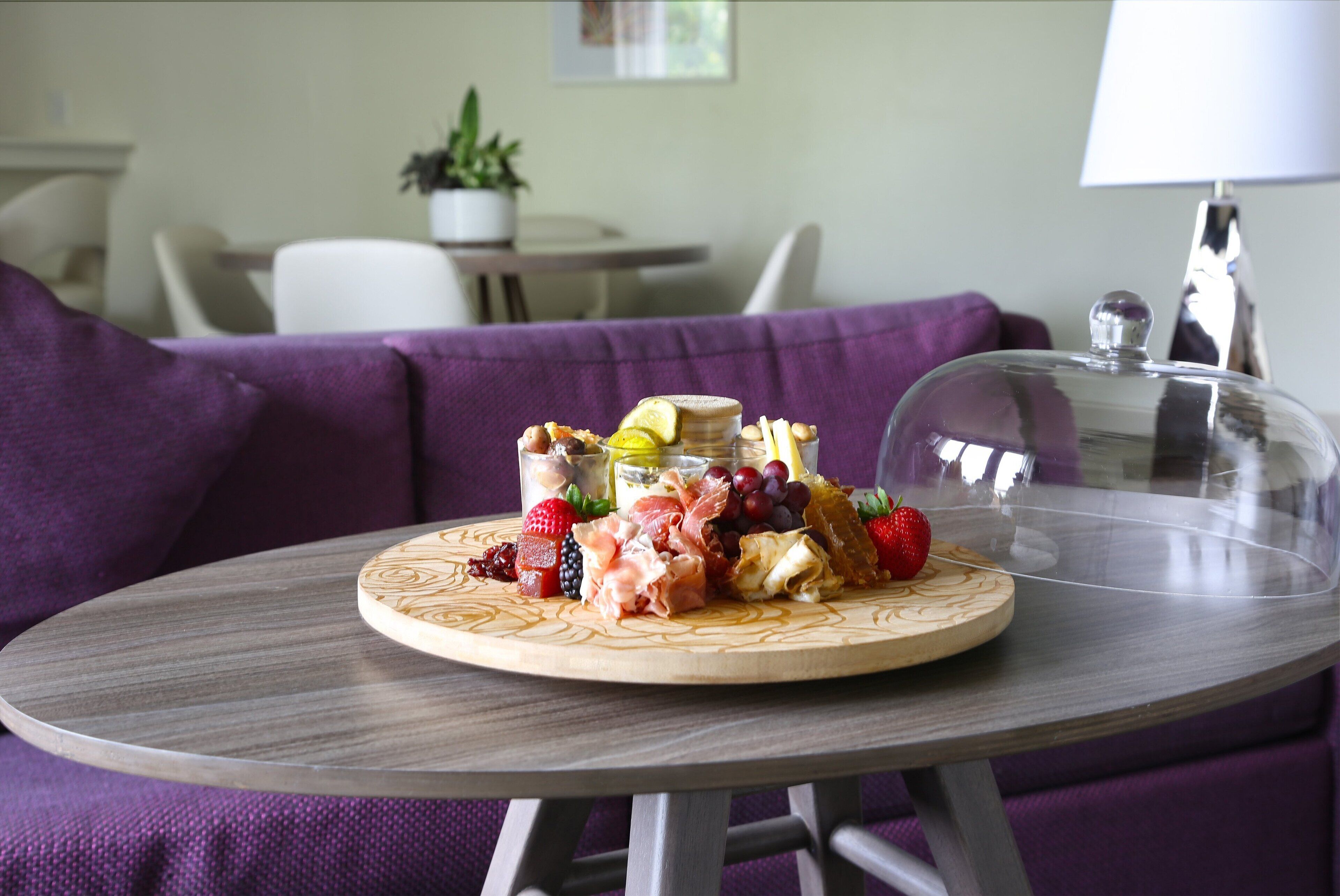 A round wooden table with a glass dome cover, displaying a charcuterie board with various meats, cheeses, and fruits.