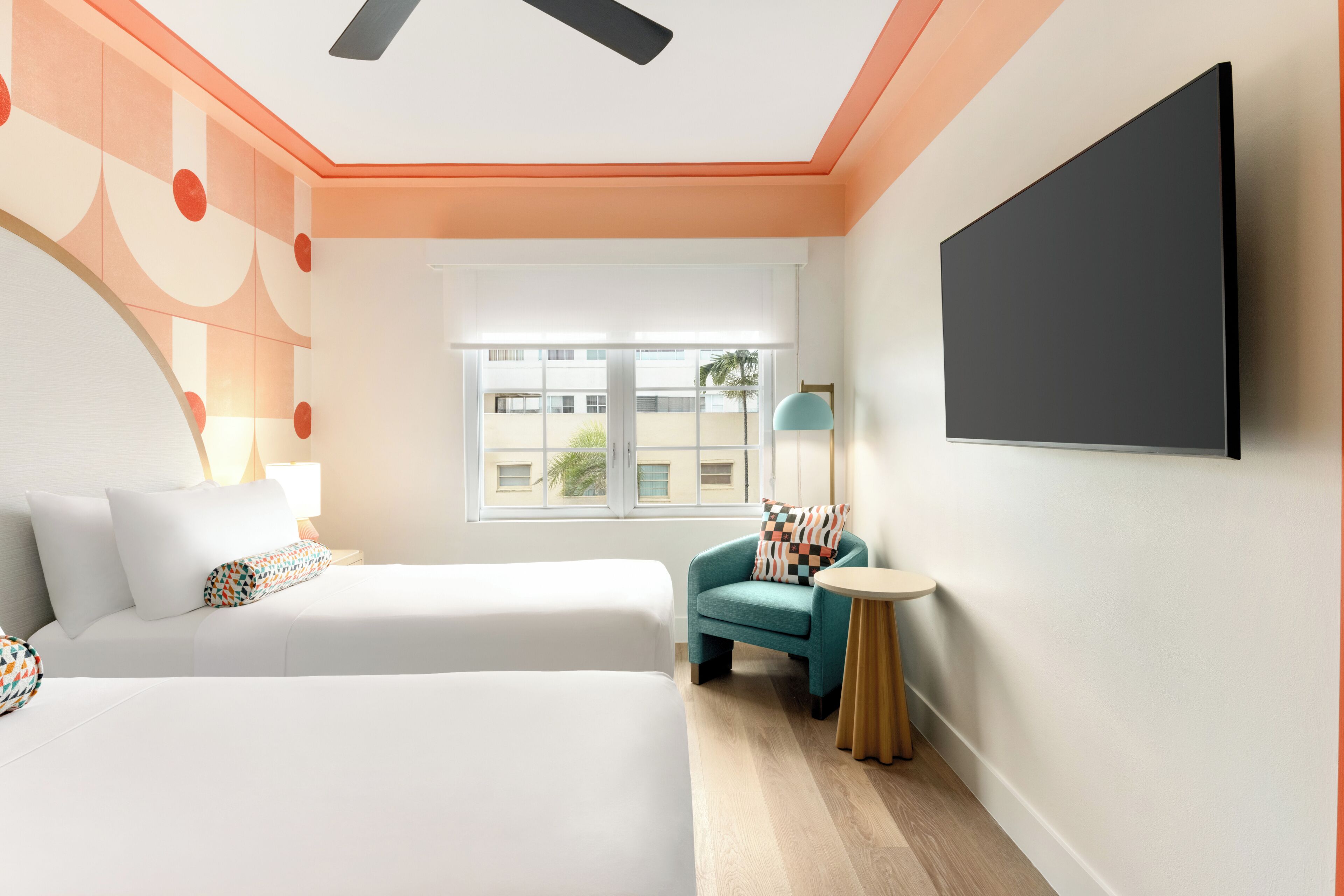 A hotel room with two beds, a teal armchair, a small round table, and a large TV on the wall.