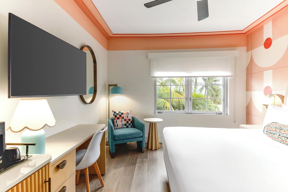 Een moderne hotelkamer met een flatscreen televisie, een bureau met lamp, een blauwgroene fauteuil, een ronde tafel en een bed met gestreepte kussens.