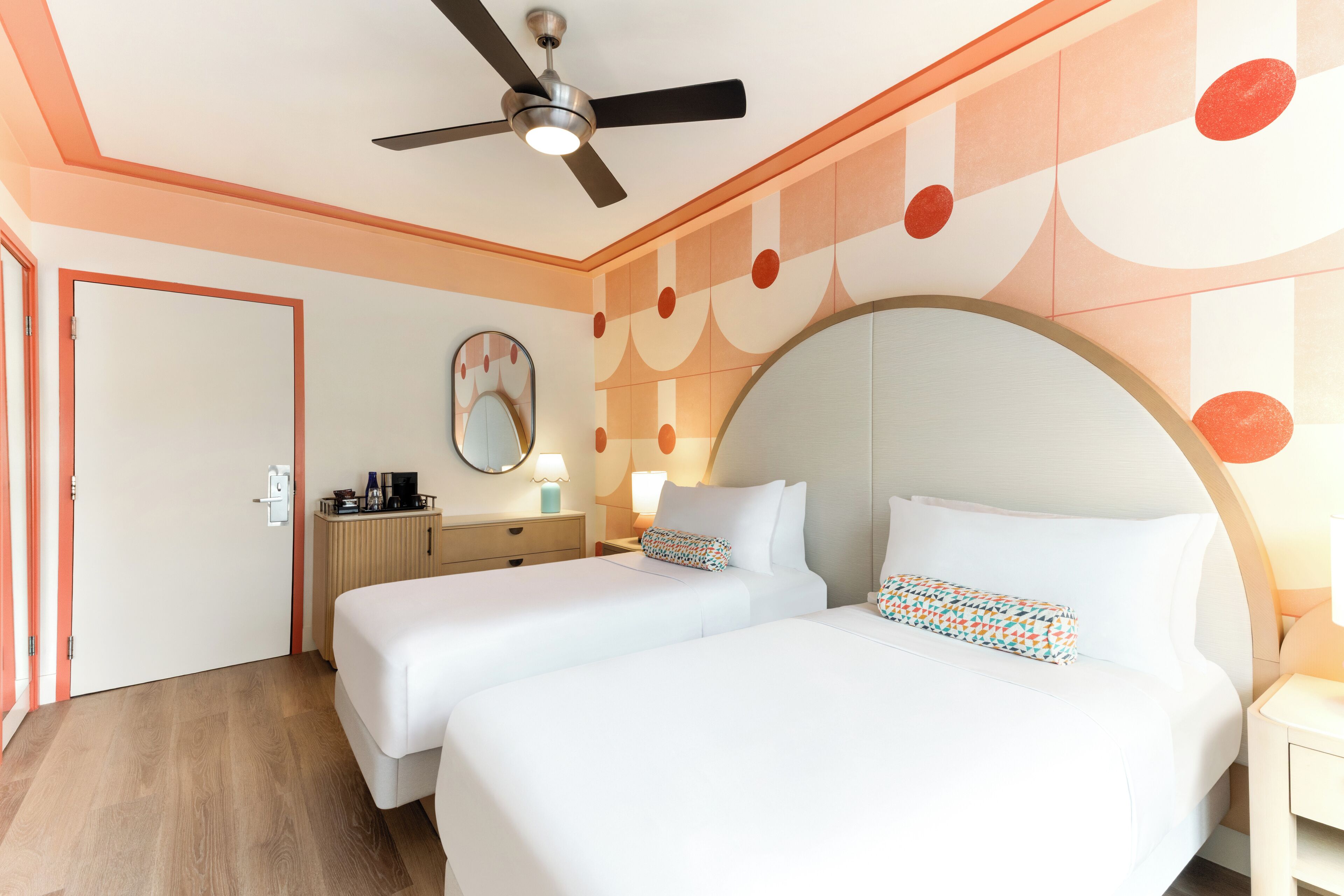 A hotel room with two beds, a ceiling fan, a nightstand with a lamp, and a wall-mounted mirror.