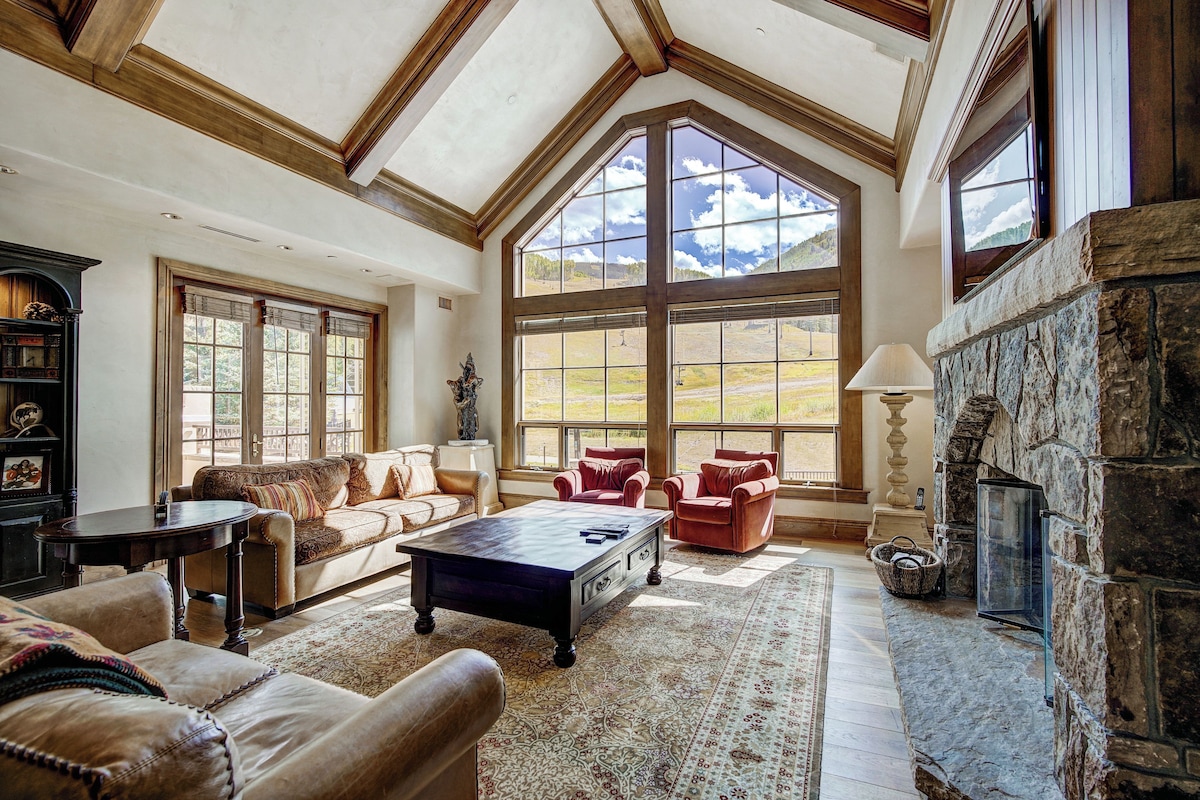 A spacious living room with a stone fireplace, large windows, and a high ceiling with wooden beams.