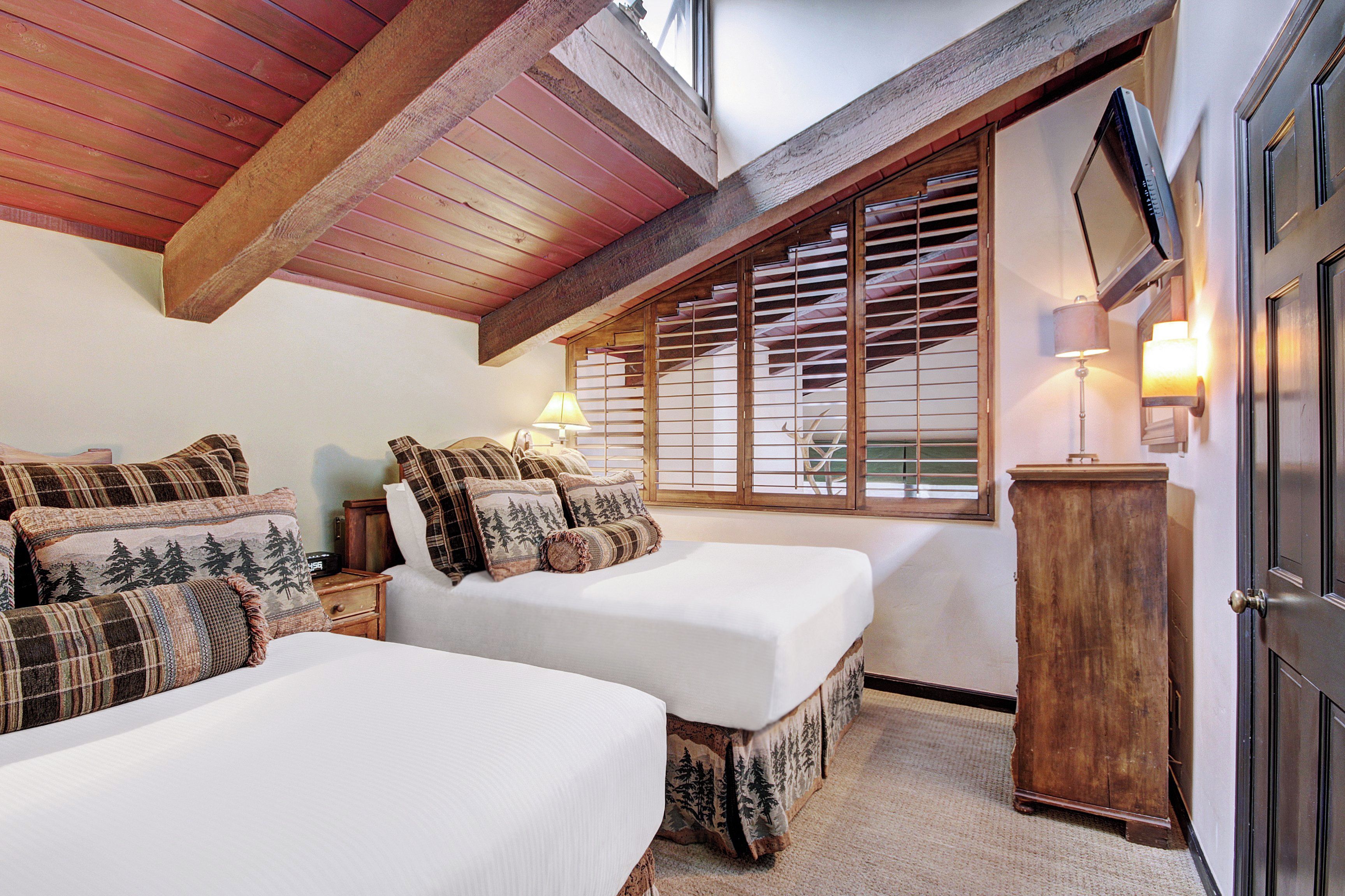 Een slaapkamer met twee bedden, houten balken, een raam met jaloezieën, een televisie en een nachtkastje.