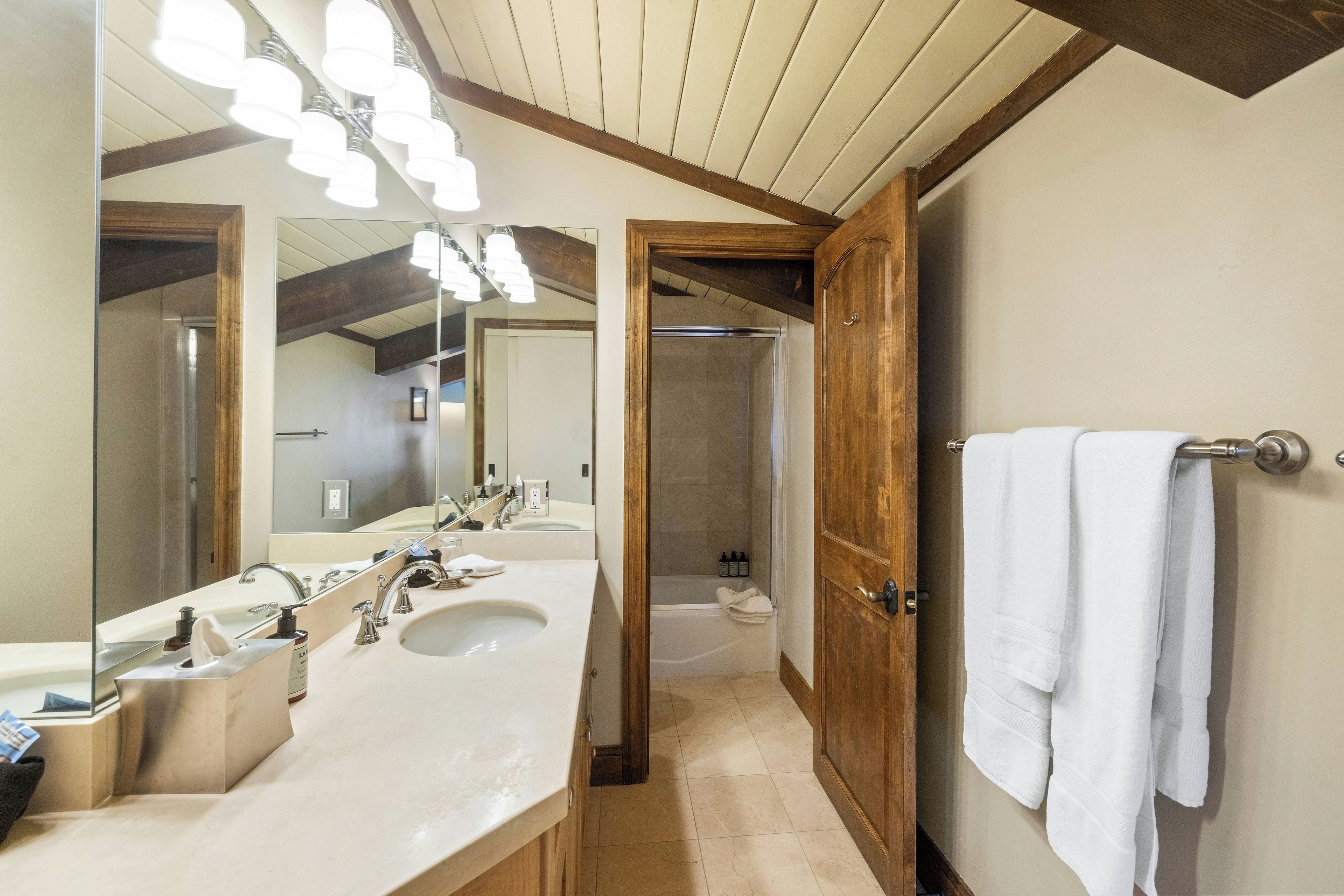 A bathroom with a double sink, a large mirror, and a wooden door leading to a shower area.