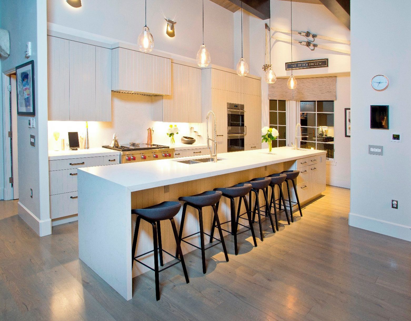 Una cocina moderna con una isla grande, taburetes de bar, iluminación colgante y un reloj visible en la pared.