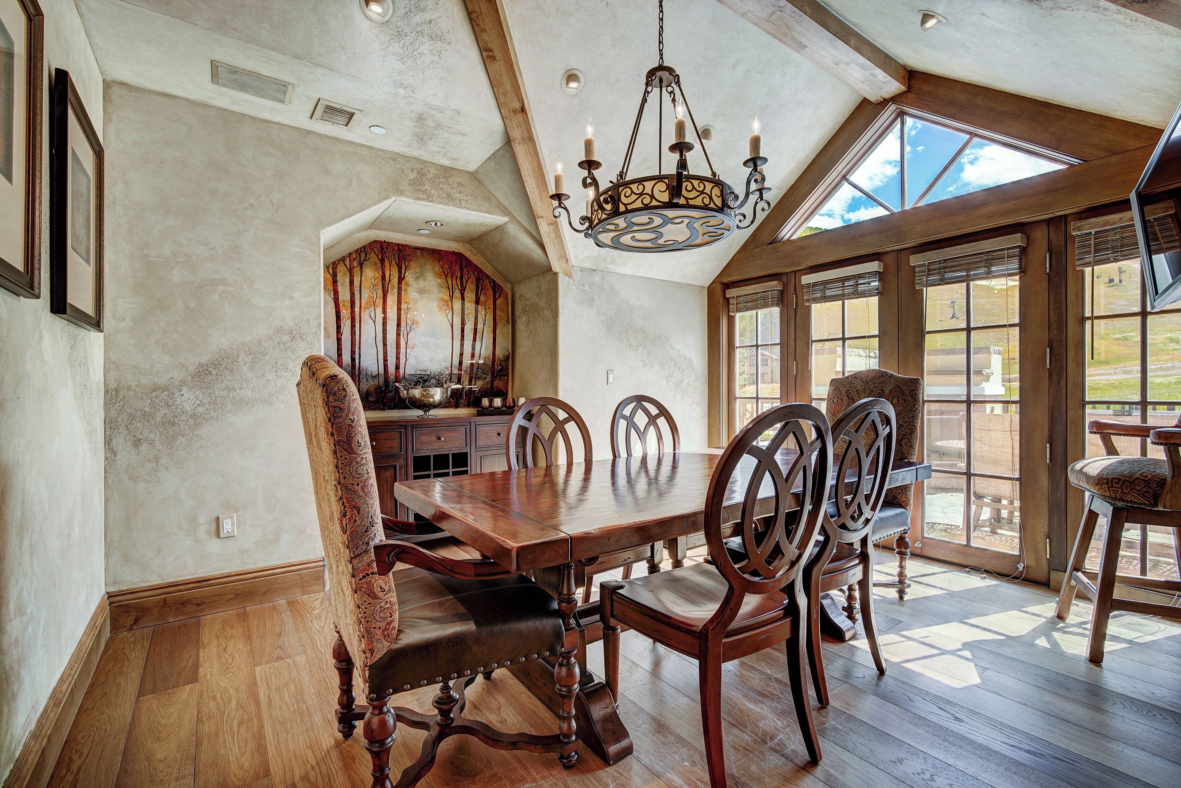 A dining area with a wooden table and chairs, a chandelier, and large windows.