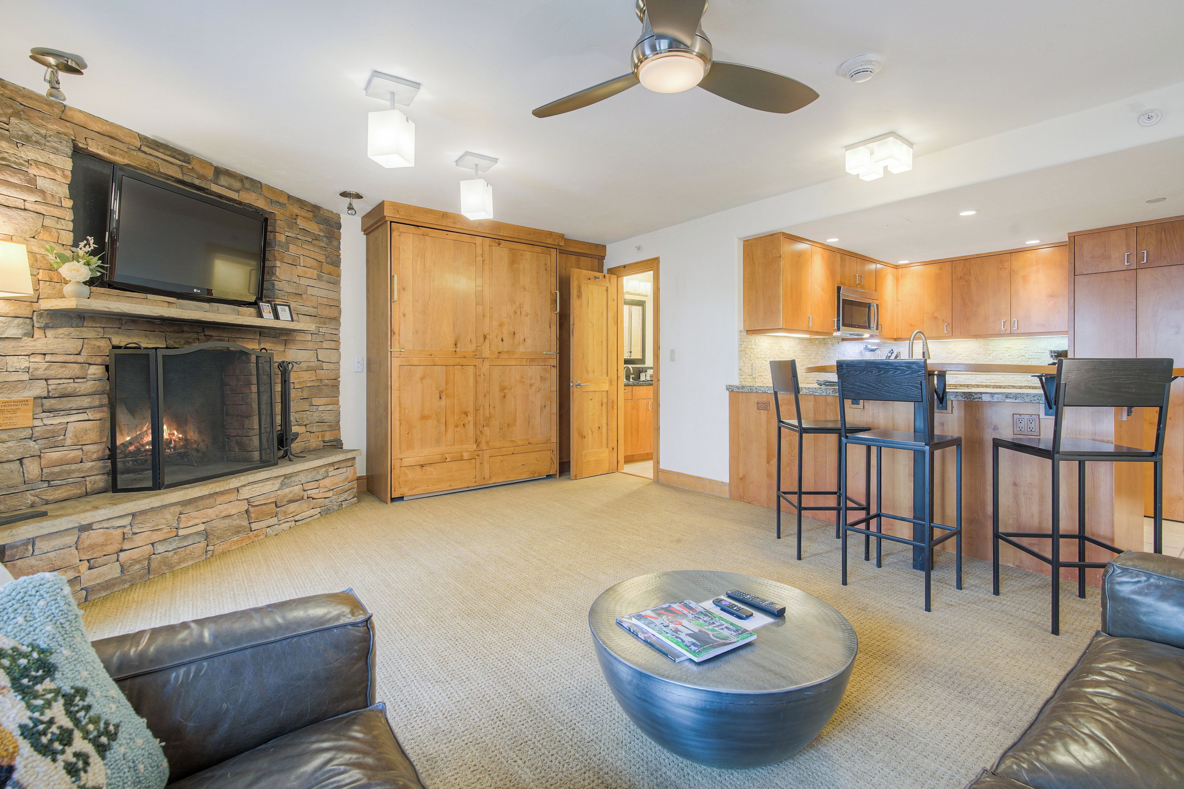 A modern living room with a stone fireplace, leather sofas, a round coffee table, and a kitchen area with wooden cabinets and bar stools.