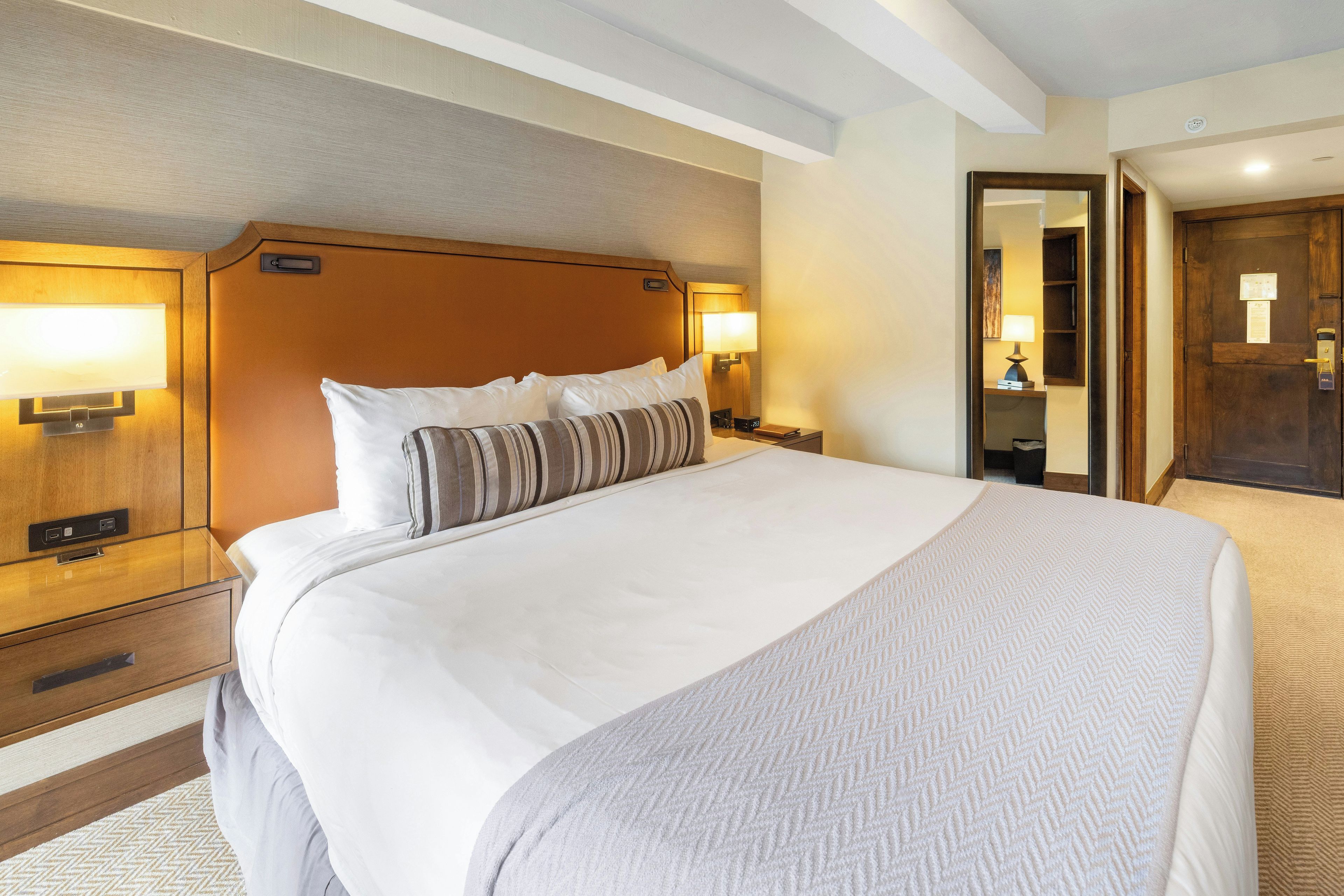 A hotel room with a large bed, two bedside tables with lamps, a wooden headboard, and a door with a mirror.