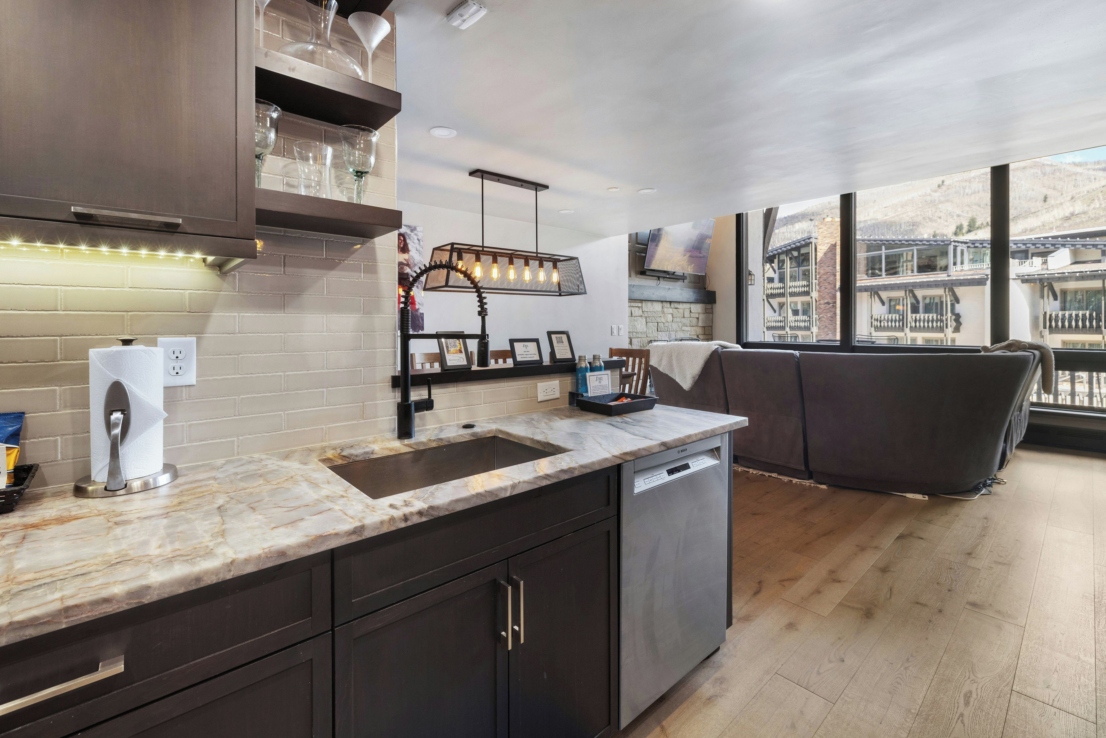 A modern kitchen with a built-in dishwasher, a marble countertop, and a large window overlooking a mountainous landscape.