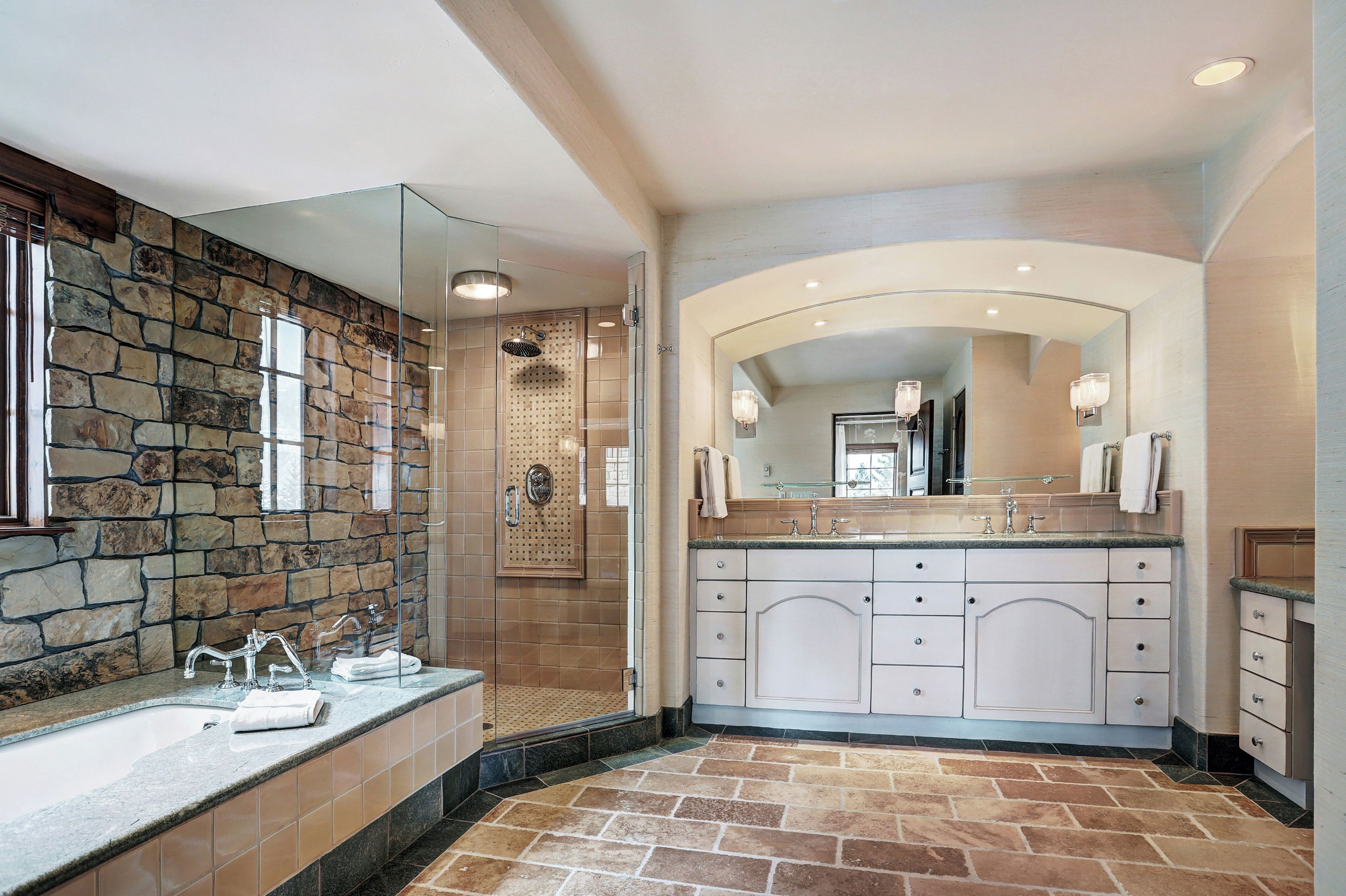 A bathroom with a stone wall, a glass shower enclosure, a large mirror, and a vanity with multiple sinks.