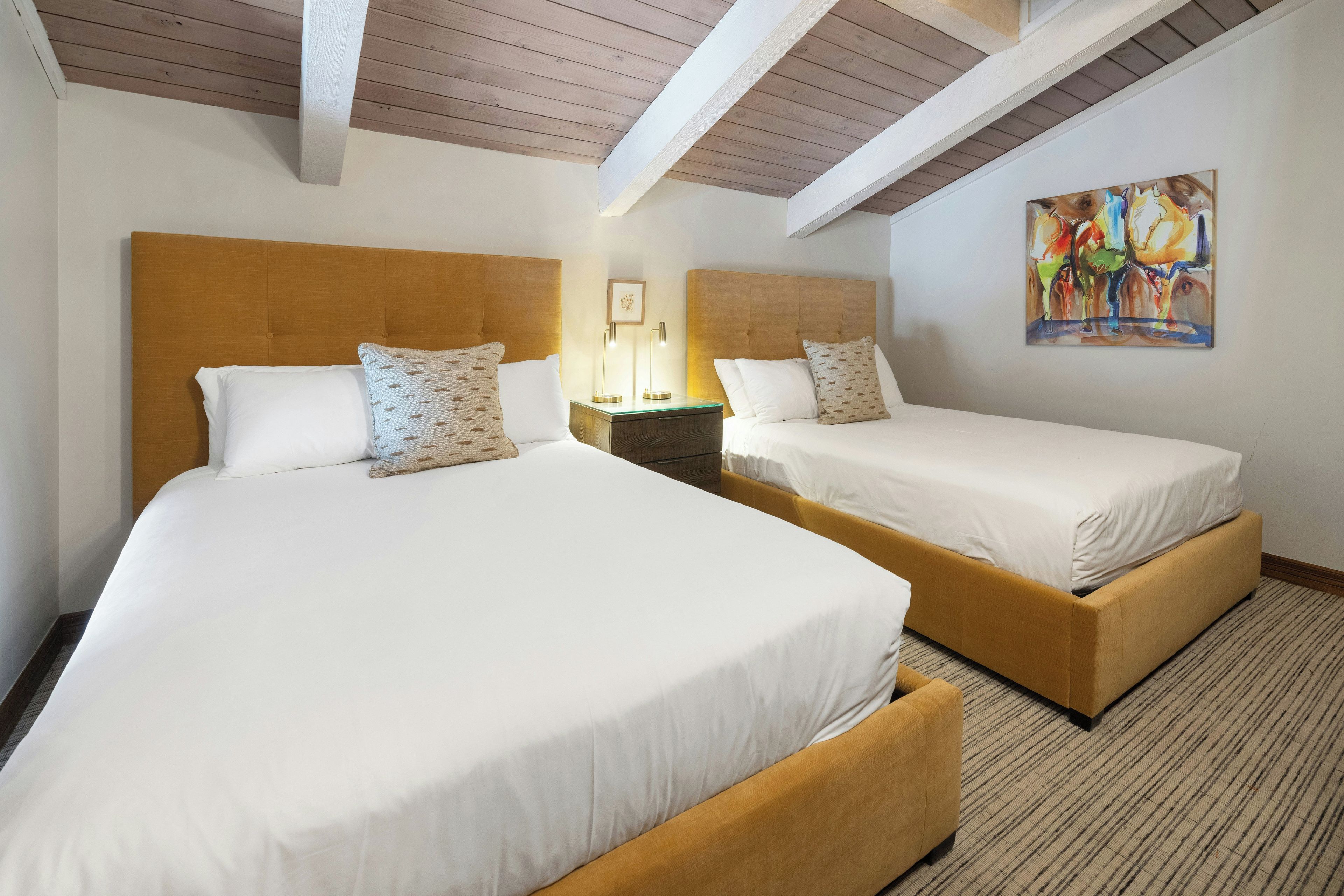 A hotel room with two beds, a nightstand, a painting, and a wooden ceiling.