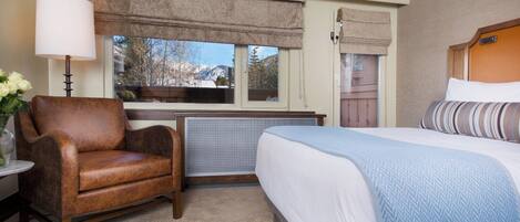 A hotel room with a bed, a chair, a lamp, and a window with a view of mountains.
