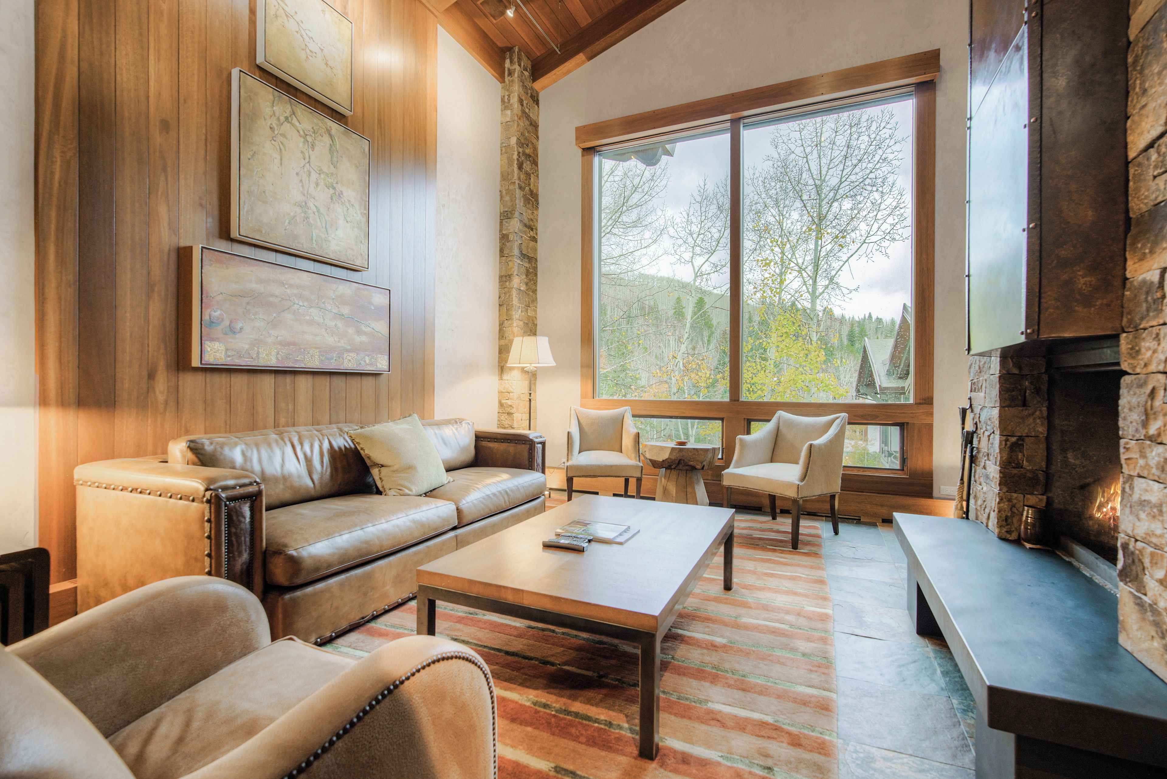 Un salon chaleureux avec un canapé en cuir, une table basse et une cheminée en pierre. De grandes fenêtres offrent une vue sur les arbres et les collines.