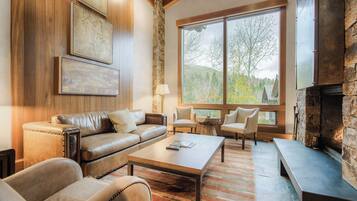 Un salon chaleureux avec un canapé en cuir, une table basse et une cheminée en pierre. De grandes fenêtres offrent une vue sur les arbres et les collines.