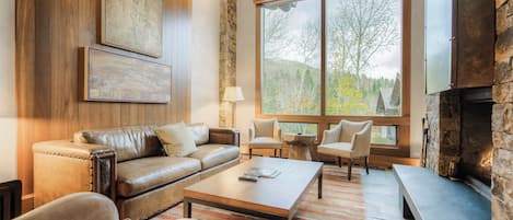 A cozy living room with a leather sofa, a coffee table, and a stone fireplace. There are large windows offering a view of trees and hills.
