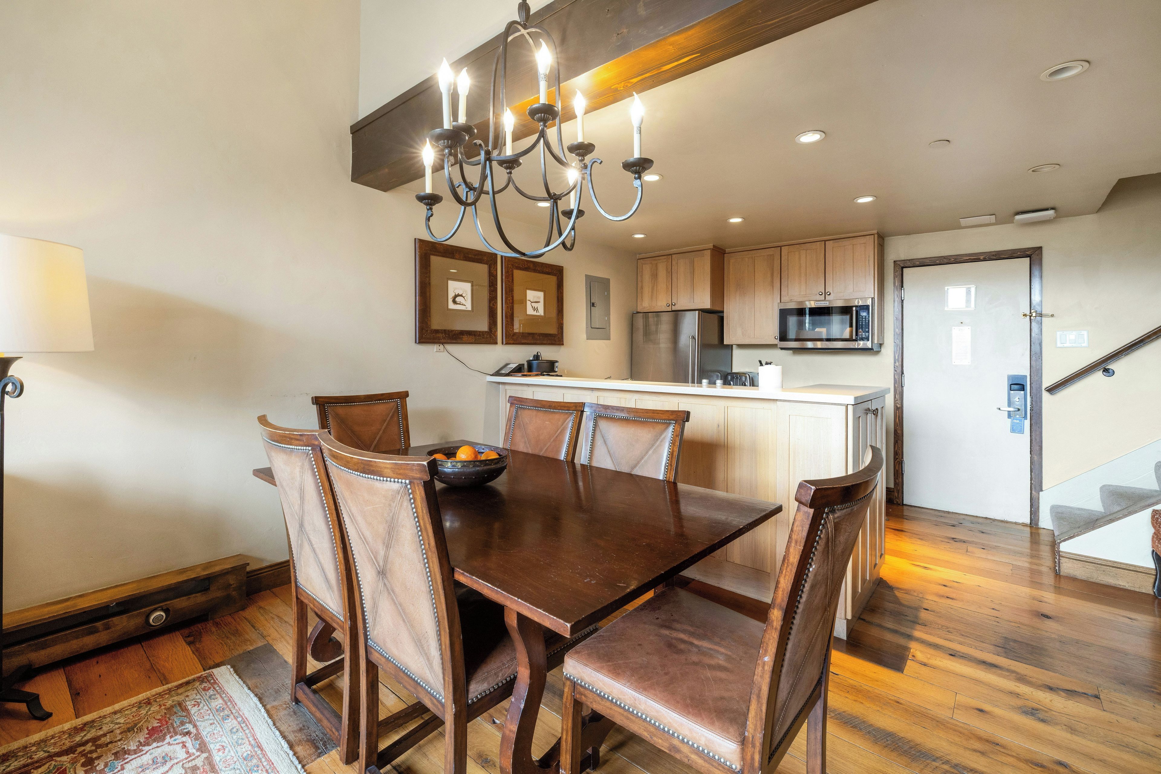A dining area with a wooden table and chairs, a chandelier, and a kitchen in the background.