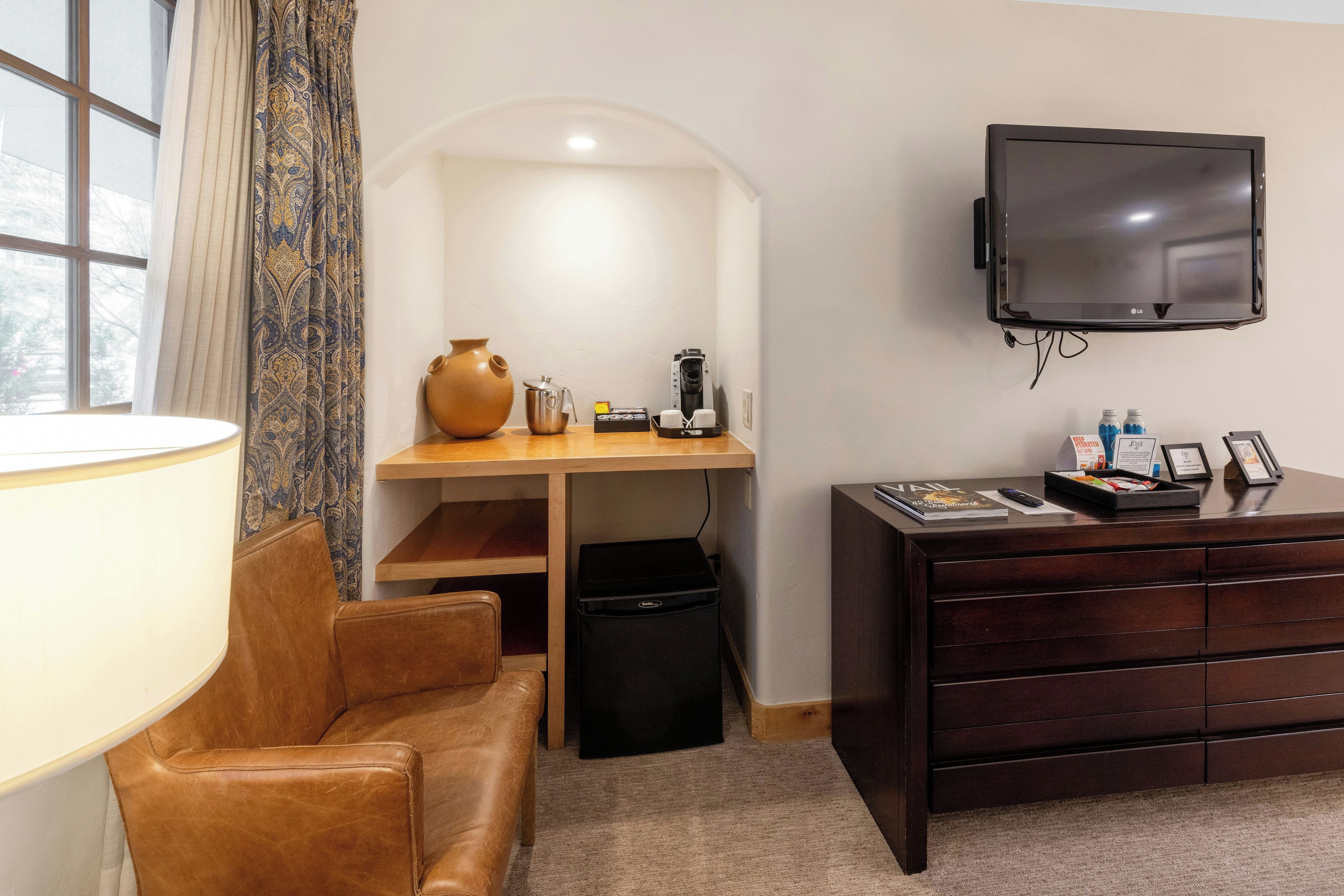 Habitación de hotel con sofá de cuero, escritorio de madera con un florero, cafetera, televisor y cómoda.