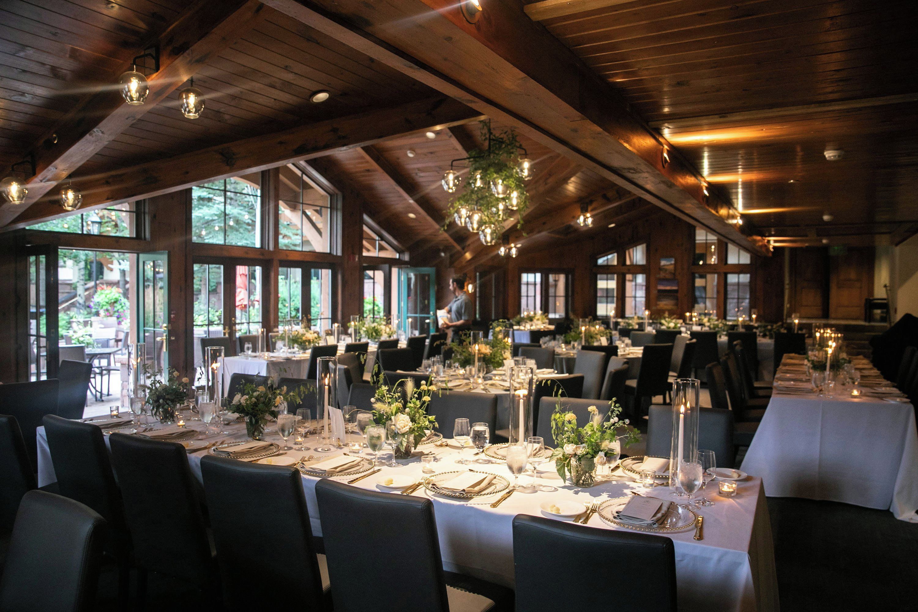 A dining area with multiple tables set for a formal event, featuring chandeliers, candles, and floral centerpieces.