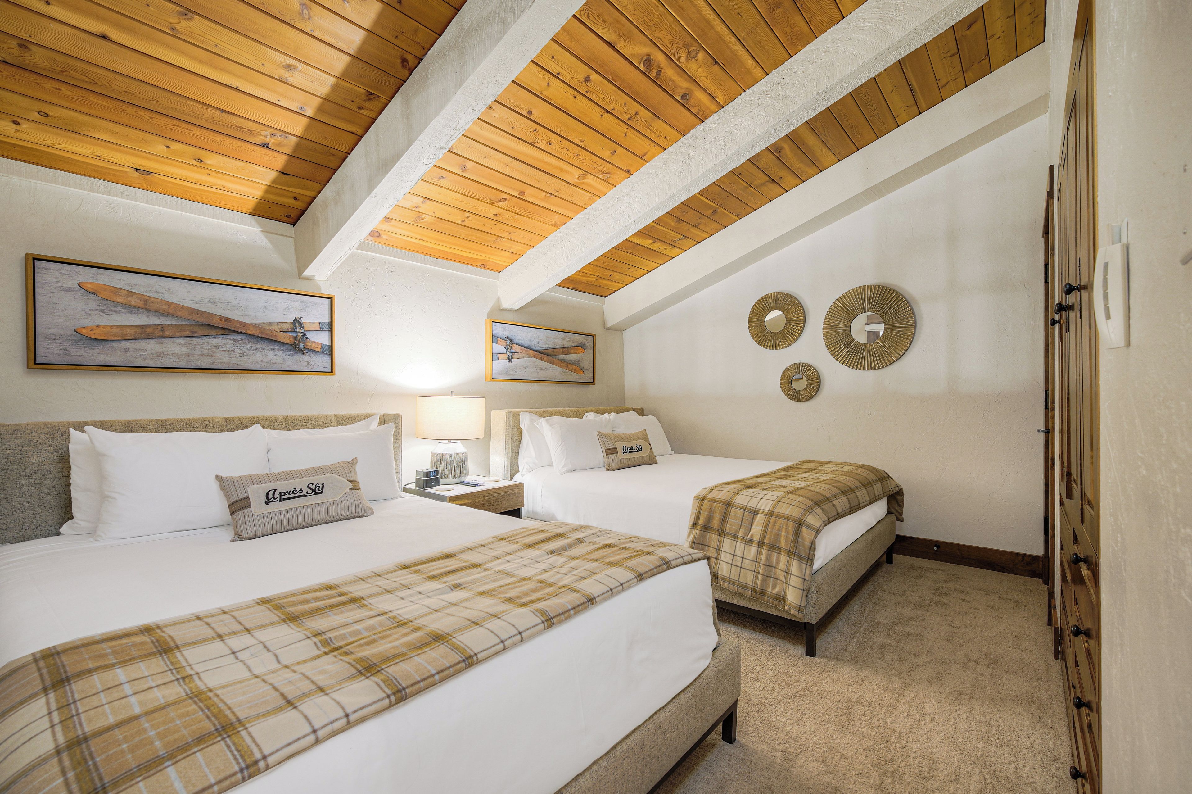 Habitación de hotel con dos camas, vigas de madera en el techo, obras de arte con temática de esquí y un reloj montado en la pared.
