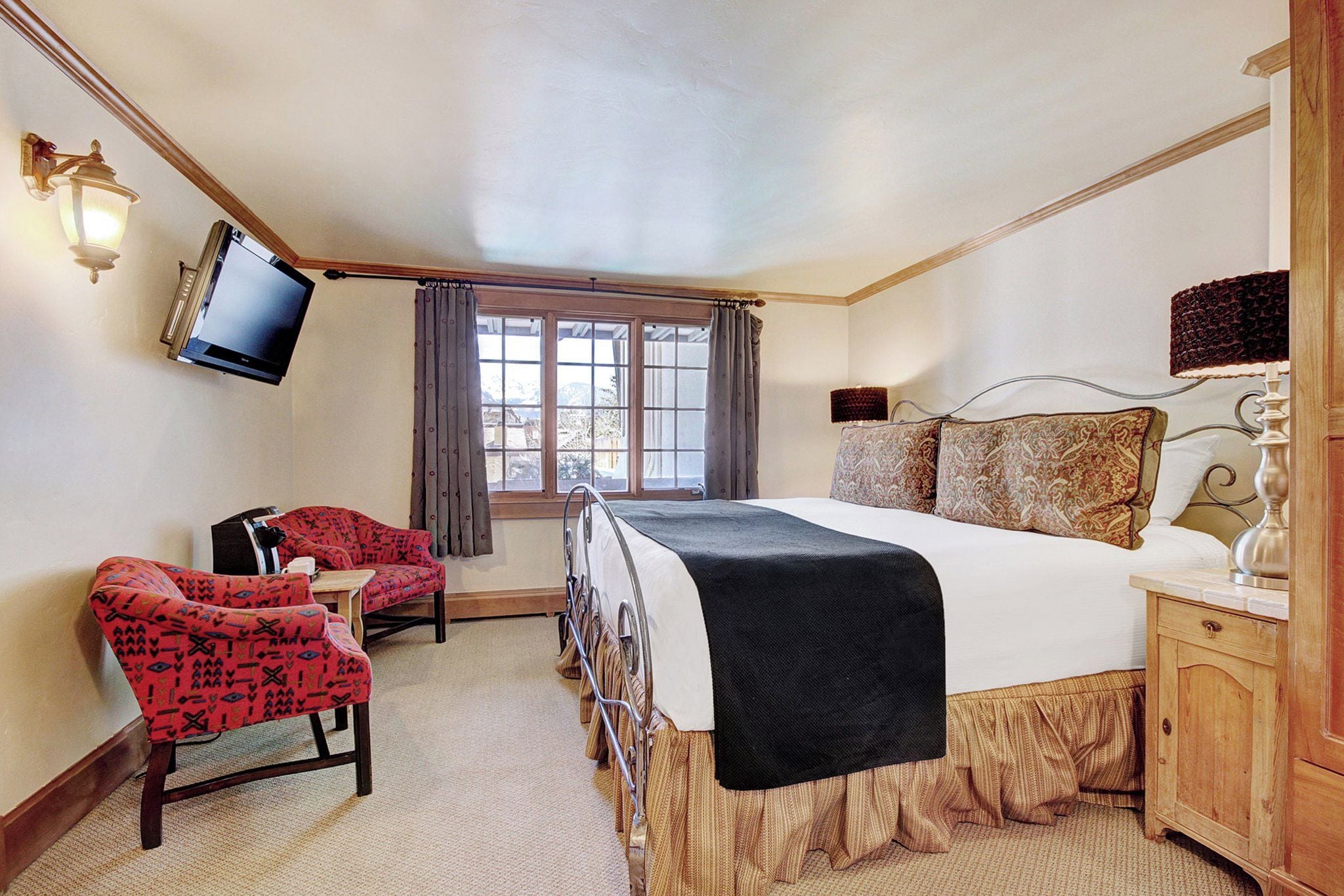 A hotel room with a bed, a TV mounted on the wall, a chair, a bedside table, and a window with curtains.