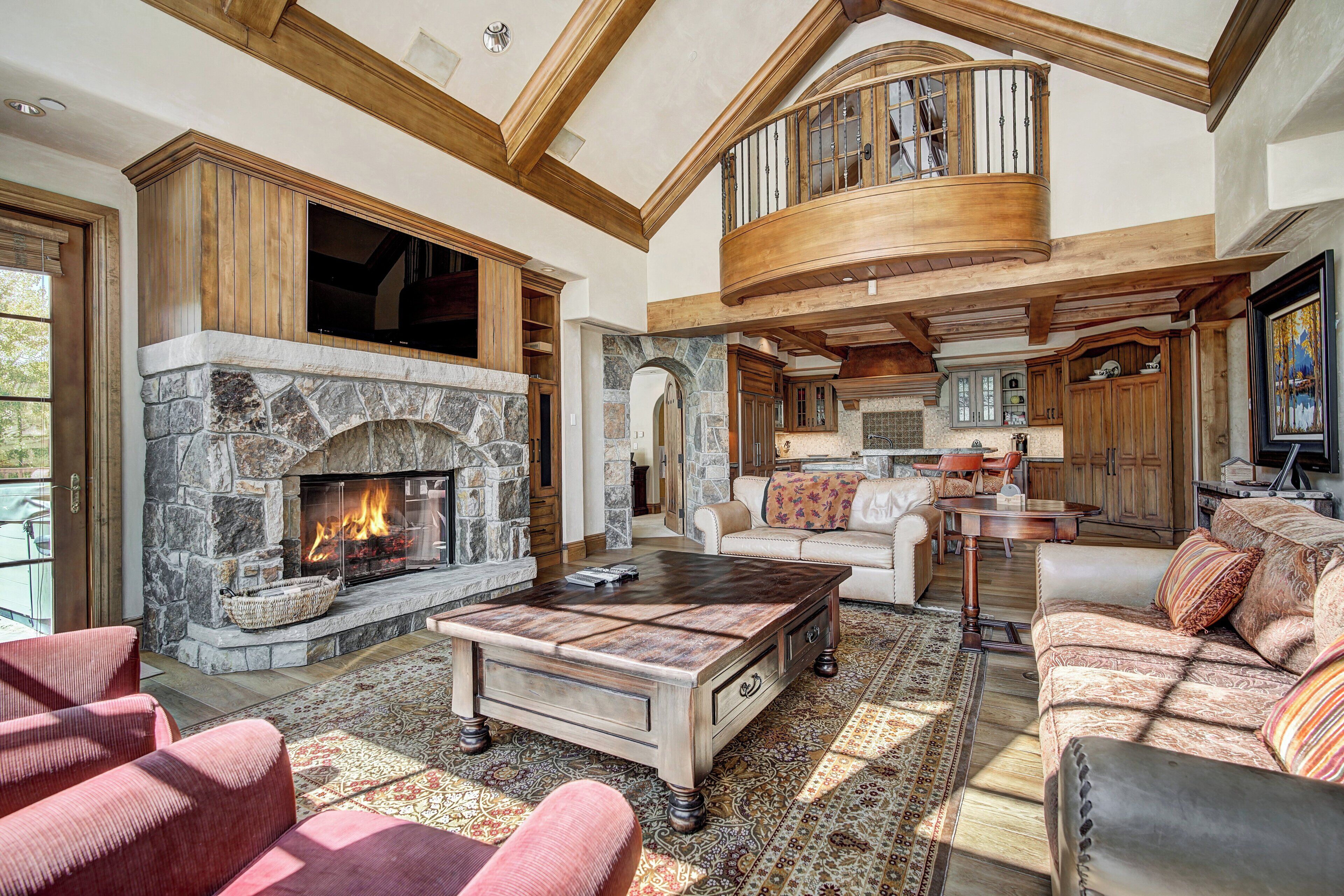 A spacious living room with a stone fireplace, wooden beams, a high ceiling, and a cozy seating area.