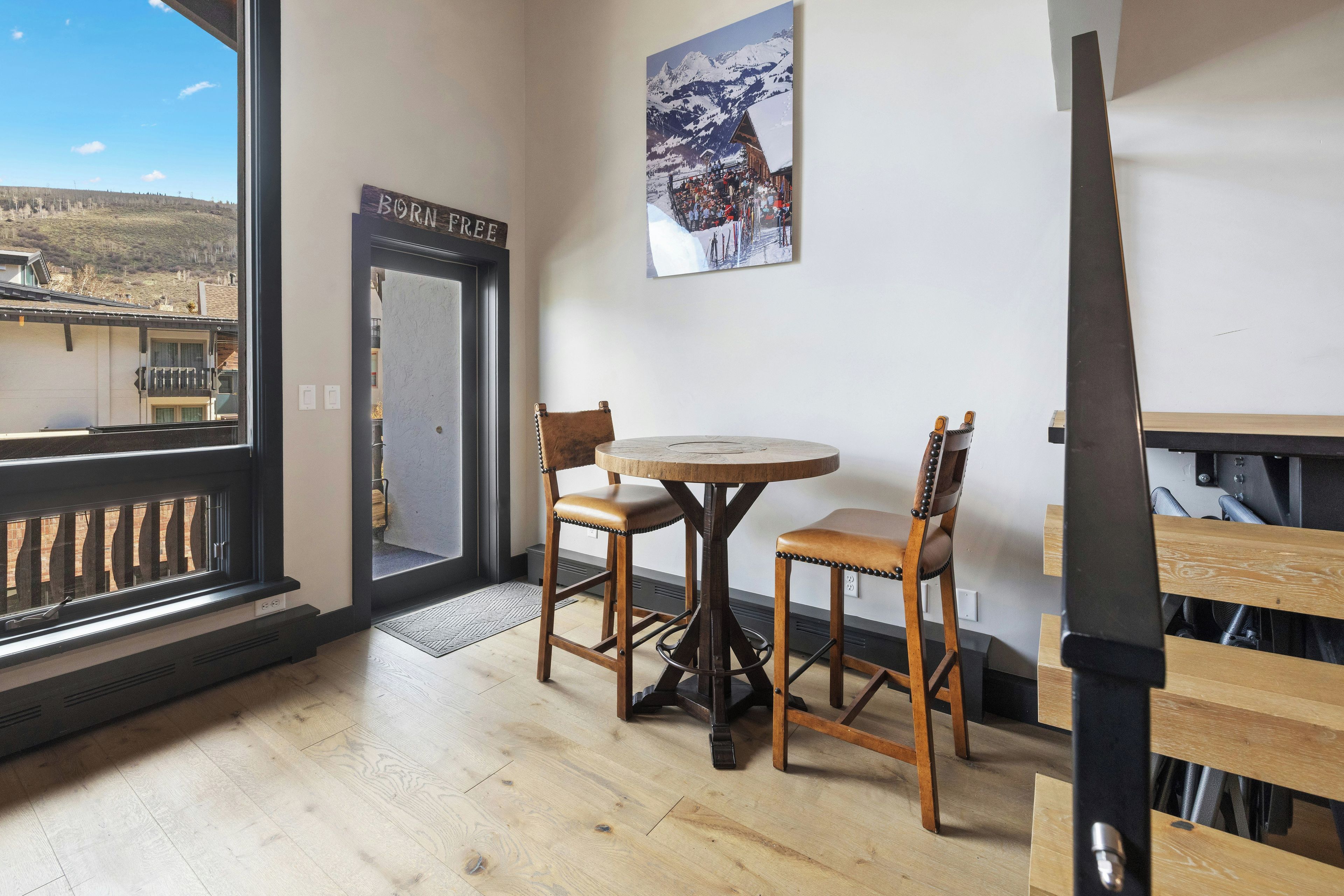 Una habitación con piso de madera, una mesa redonda con sillas, una puerta con la leyenda 'BORN FREE' y una ventana que ofrece vistas a una montaña.