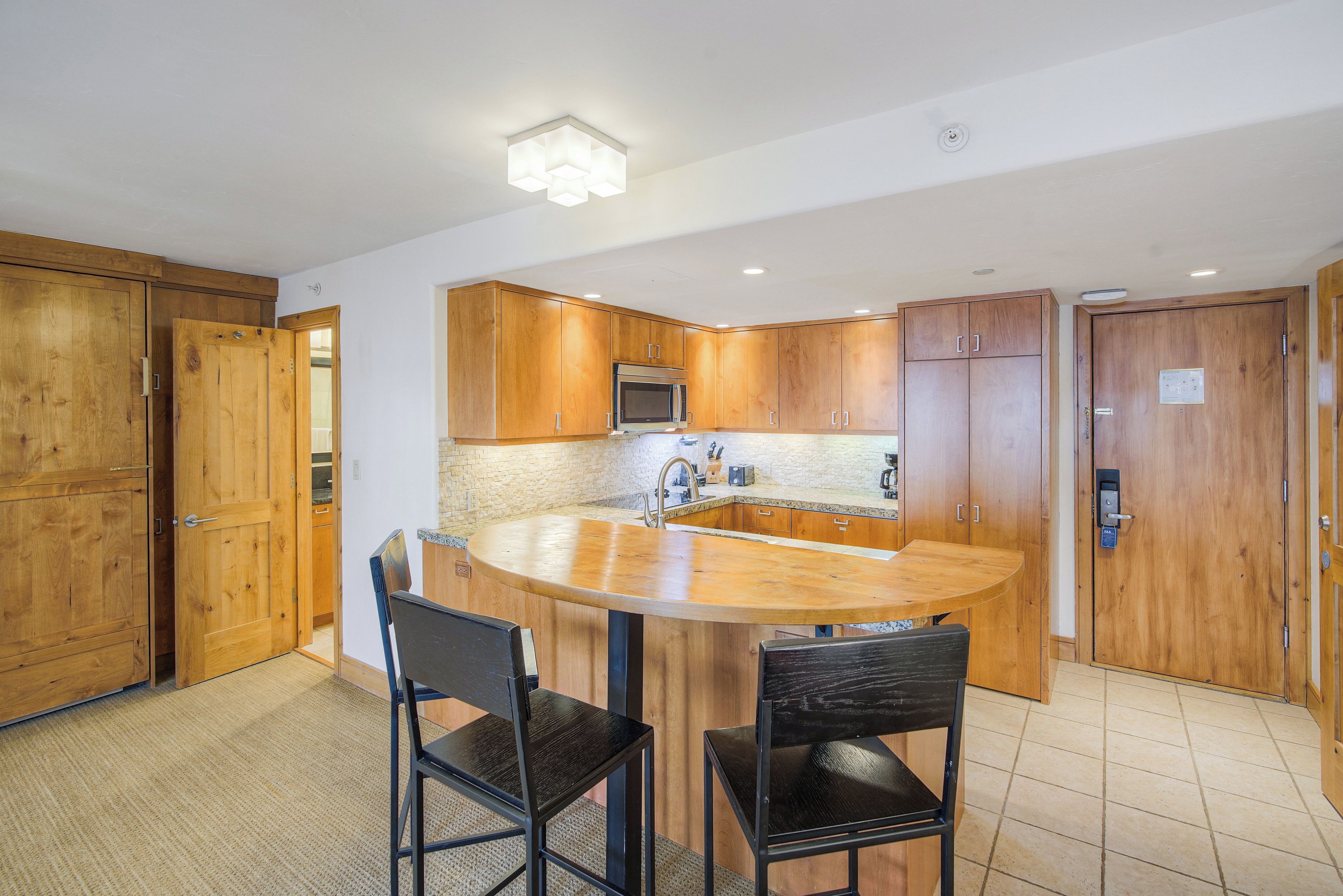 A compact kitchen with wooden cabinets, a microwave, and a sink. There are two chairs at a small island counter.