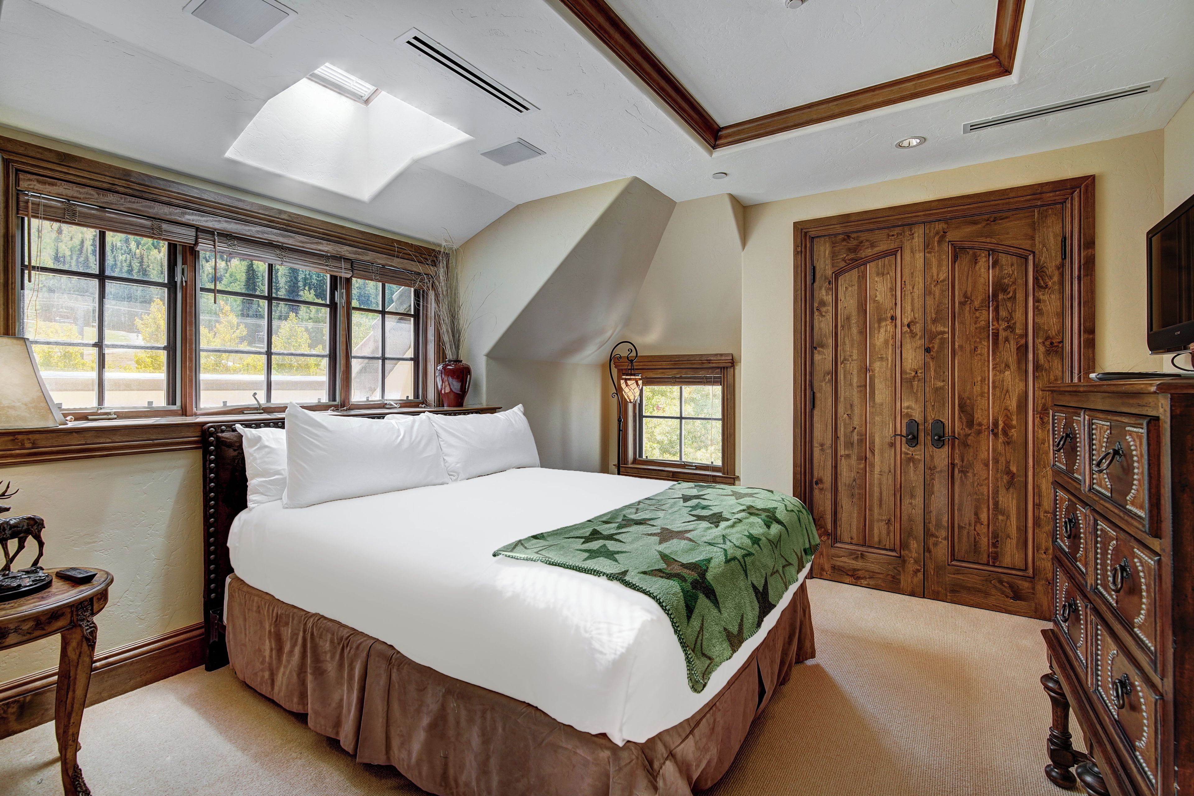 A bedroom with a large bed, a skylight, a wooden wardrobe, and a window with a view of trees.