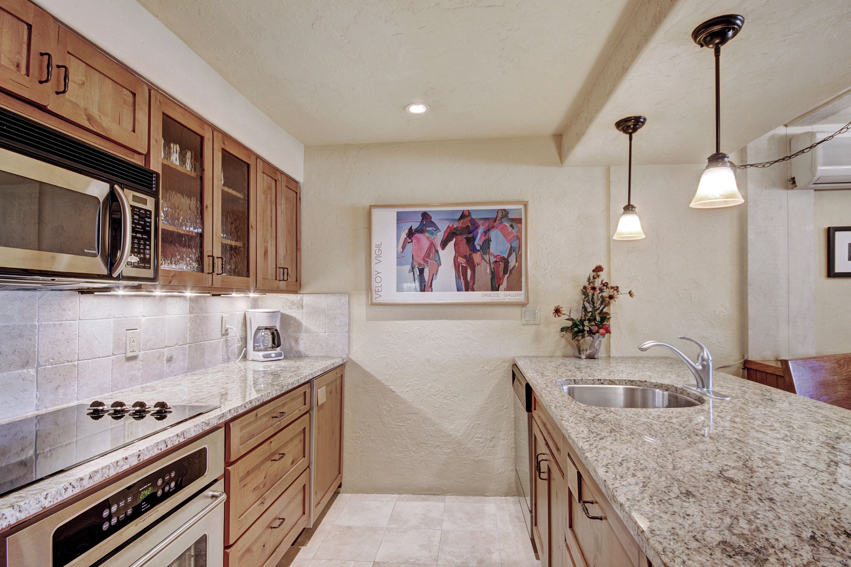 A kitchen with wooden cabinets, a stainless steel microwave, a built-in oven, granite countertops, a sink, and pendant lighting.