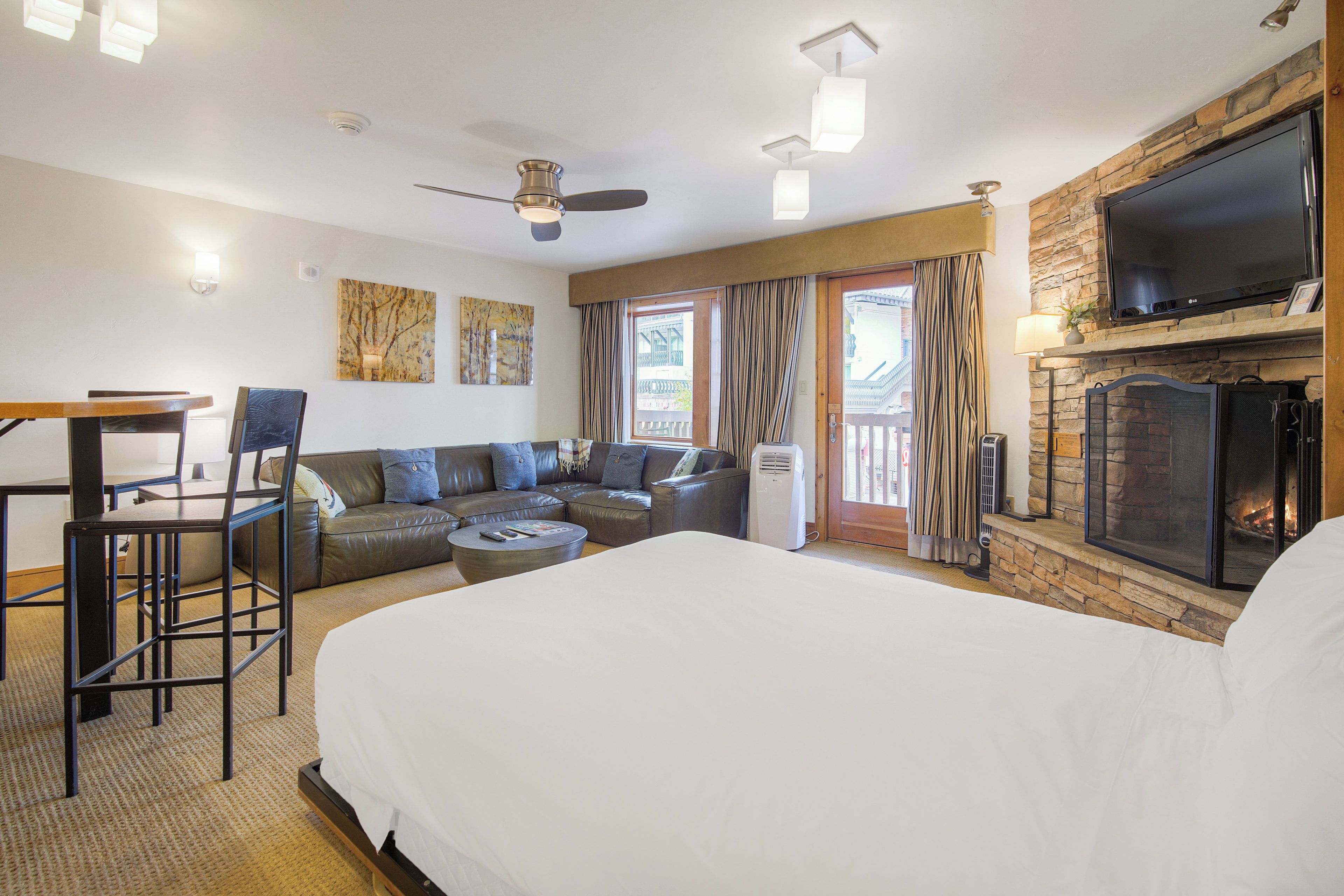 A hotel room with a large bed, a sofa, a dining table with chairs, a fireplace, and a TV mounted on the wall.