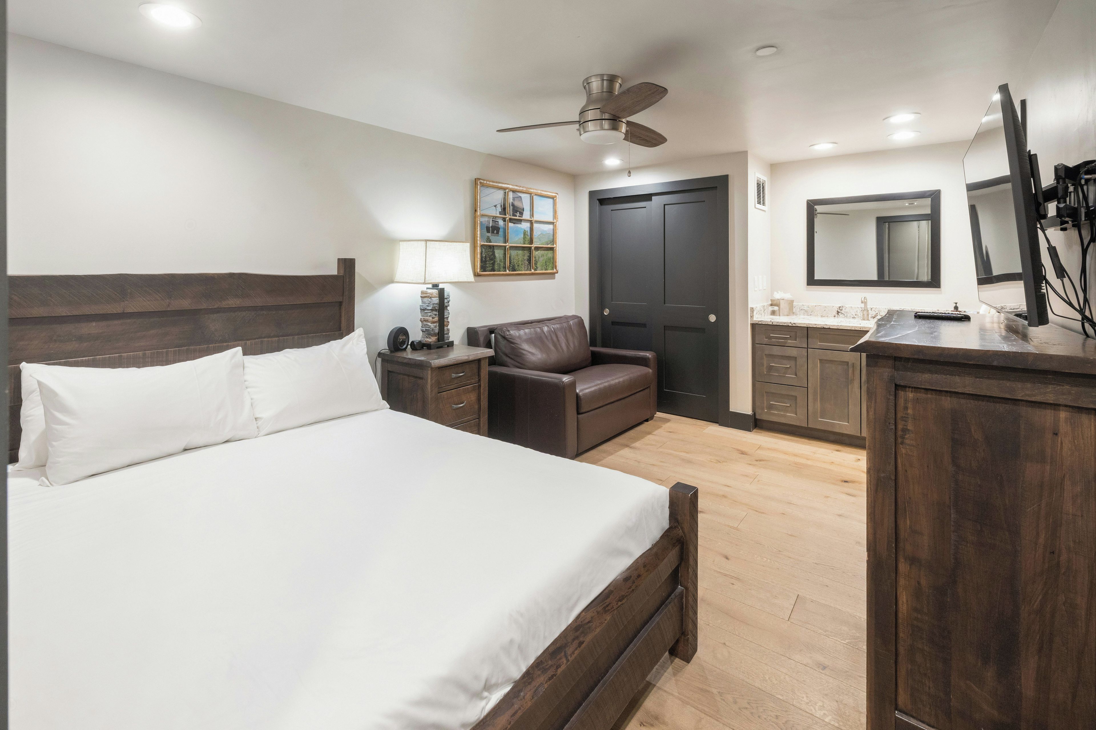 A hotel room with a large bed, a desk with a chair, a small sofa, a ceiling fan, a door, a window with a view, and a bathroom with a mirror and sink.