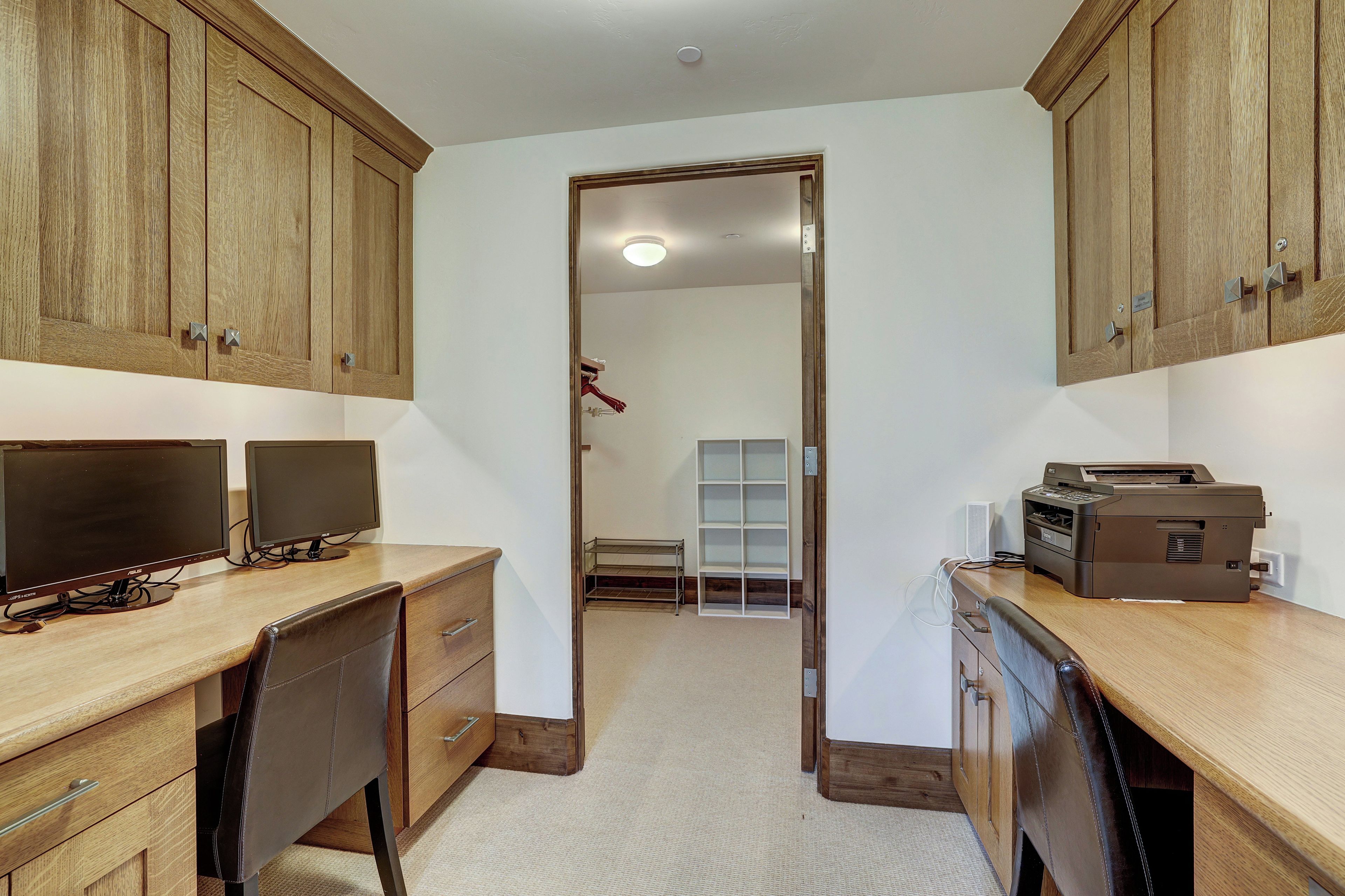 A room with a desk, two monitors, a printer, and wooden cabinets.