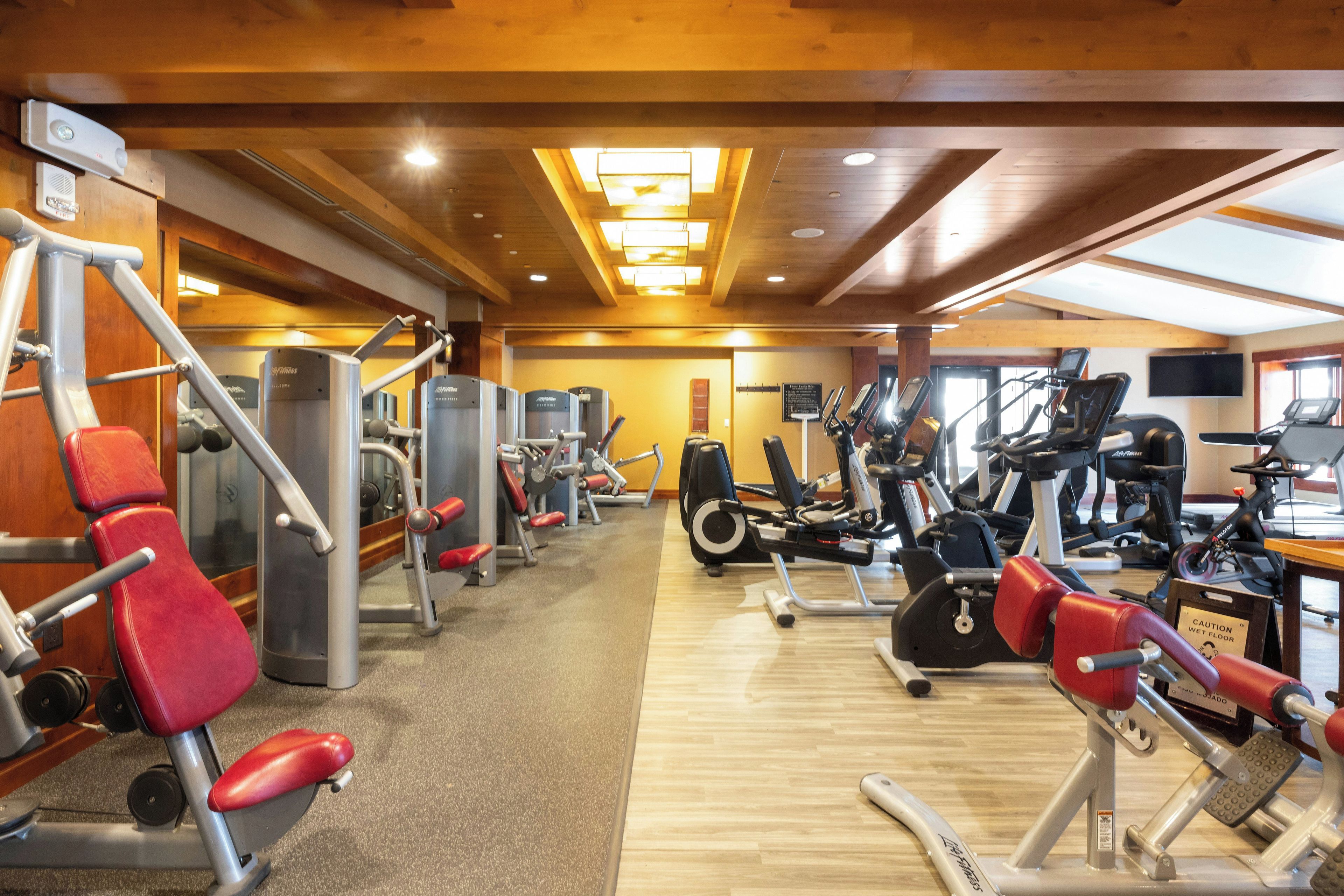 A gym with various exercise machines, wooden ceiling beams, and a caution sign on the floor.