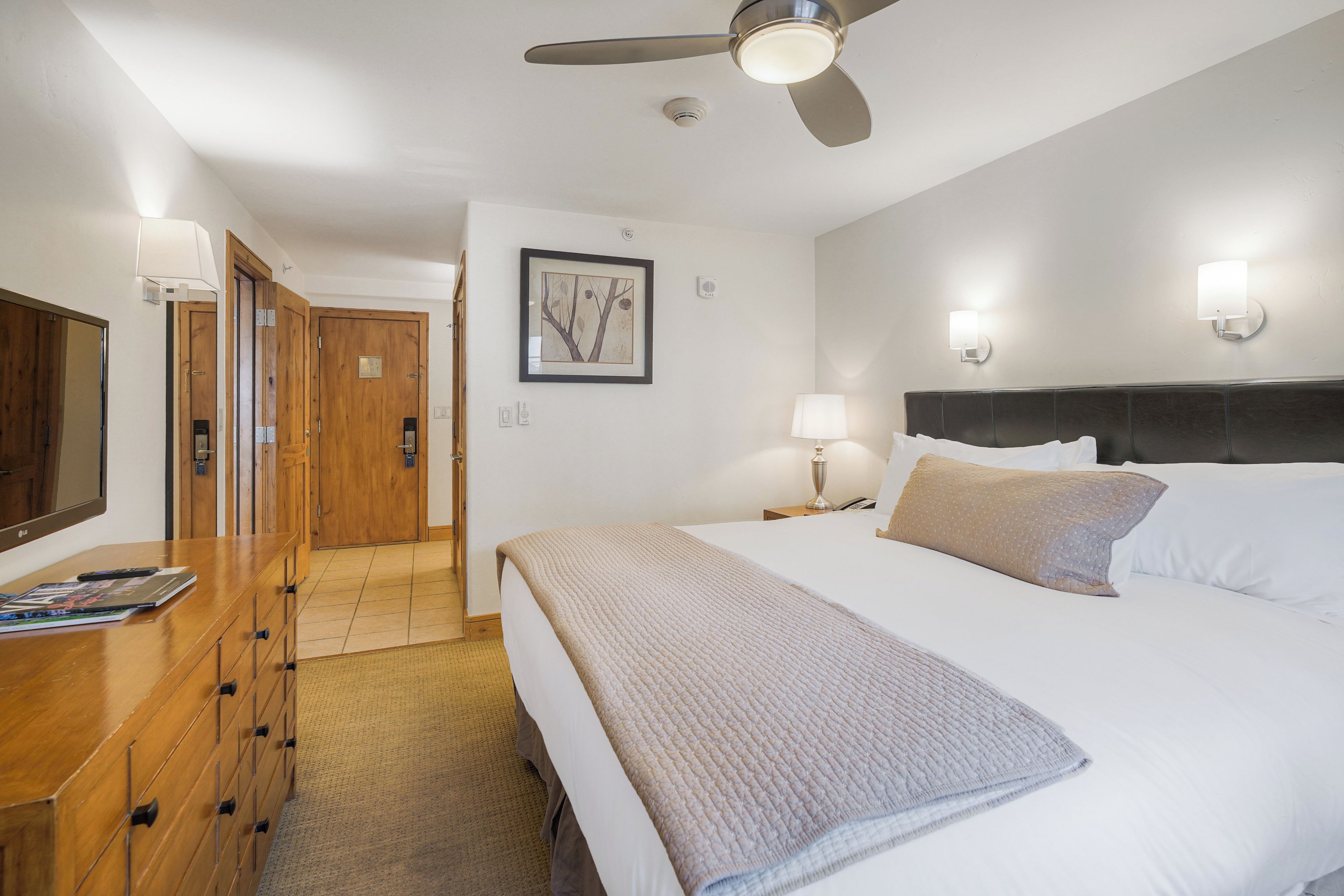 A hotel room with a bed, a wooden dresser, a ceiling fan, and a framed picture on the wall.