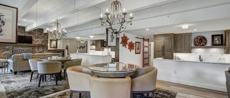 A spacious living area with a round glass table, comfortable seating, a chandelier, and a kitchen in the background.