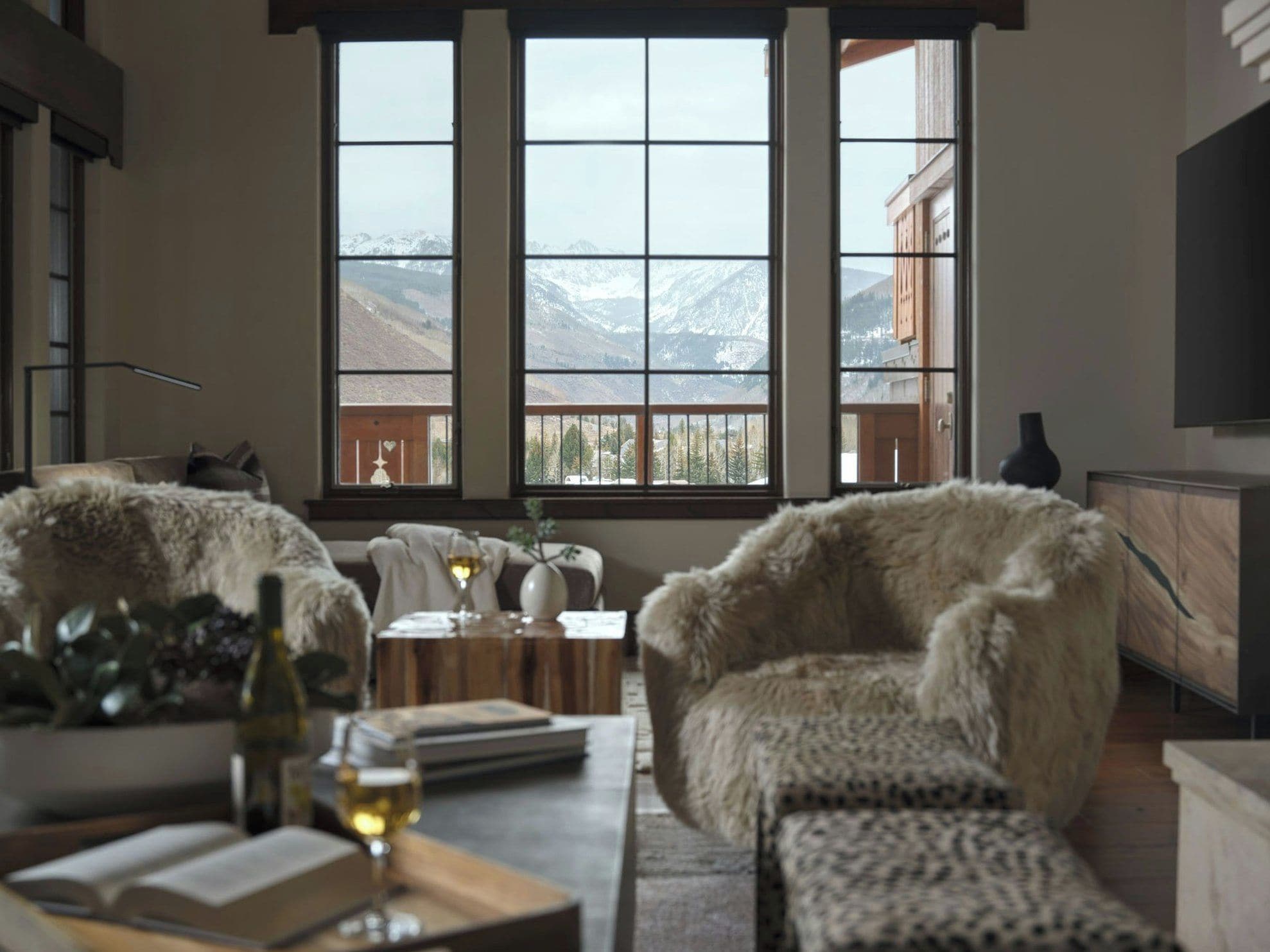 A room with a large window offering a mountain view, a sofa, a coffee table with a wine glass, and a television.