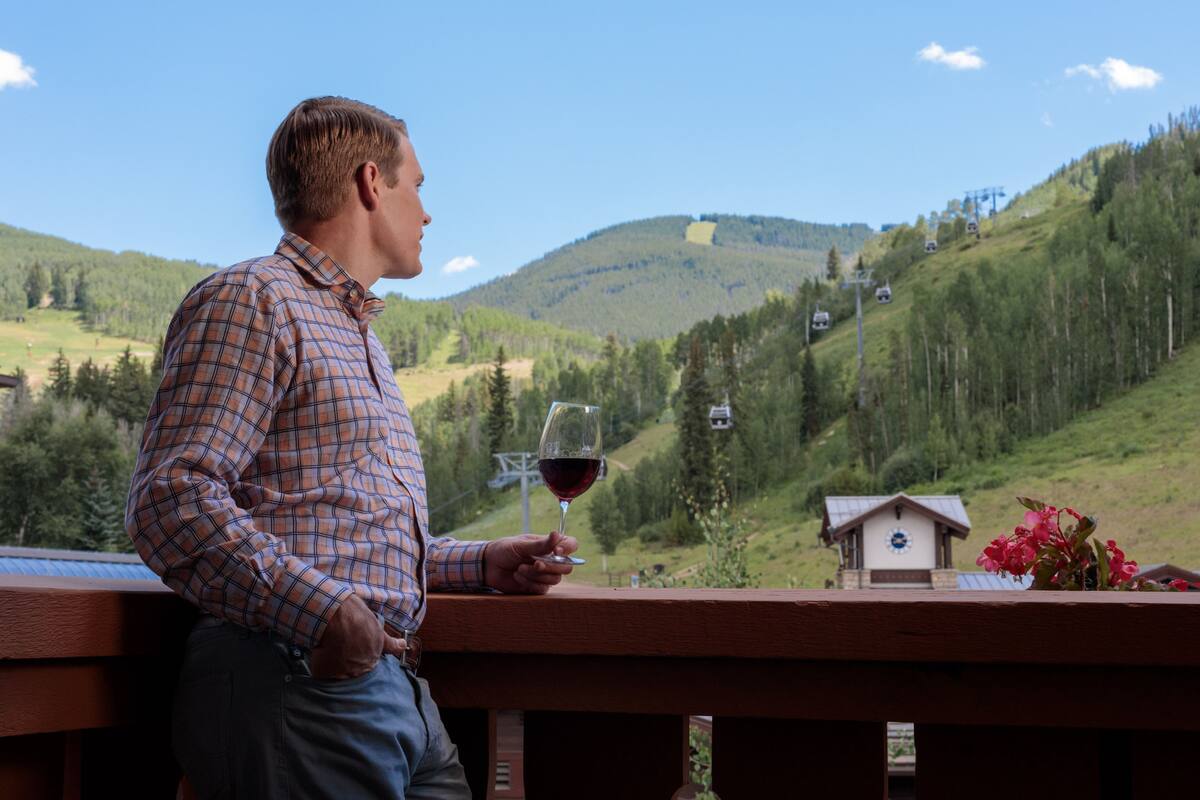 Een man staat op een balkon met uitzicht op een bergachtig landschap, met een klein huisje en een skilift op de achtergrond.