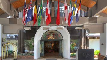 A hotel entrance with multiple national flags, a reception desk, and a bellhop cart.