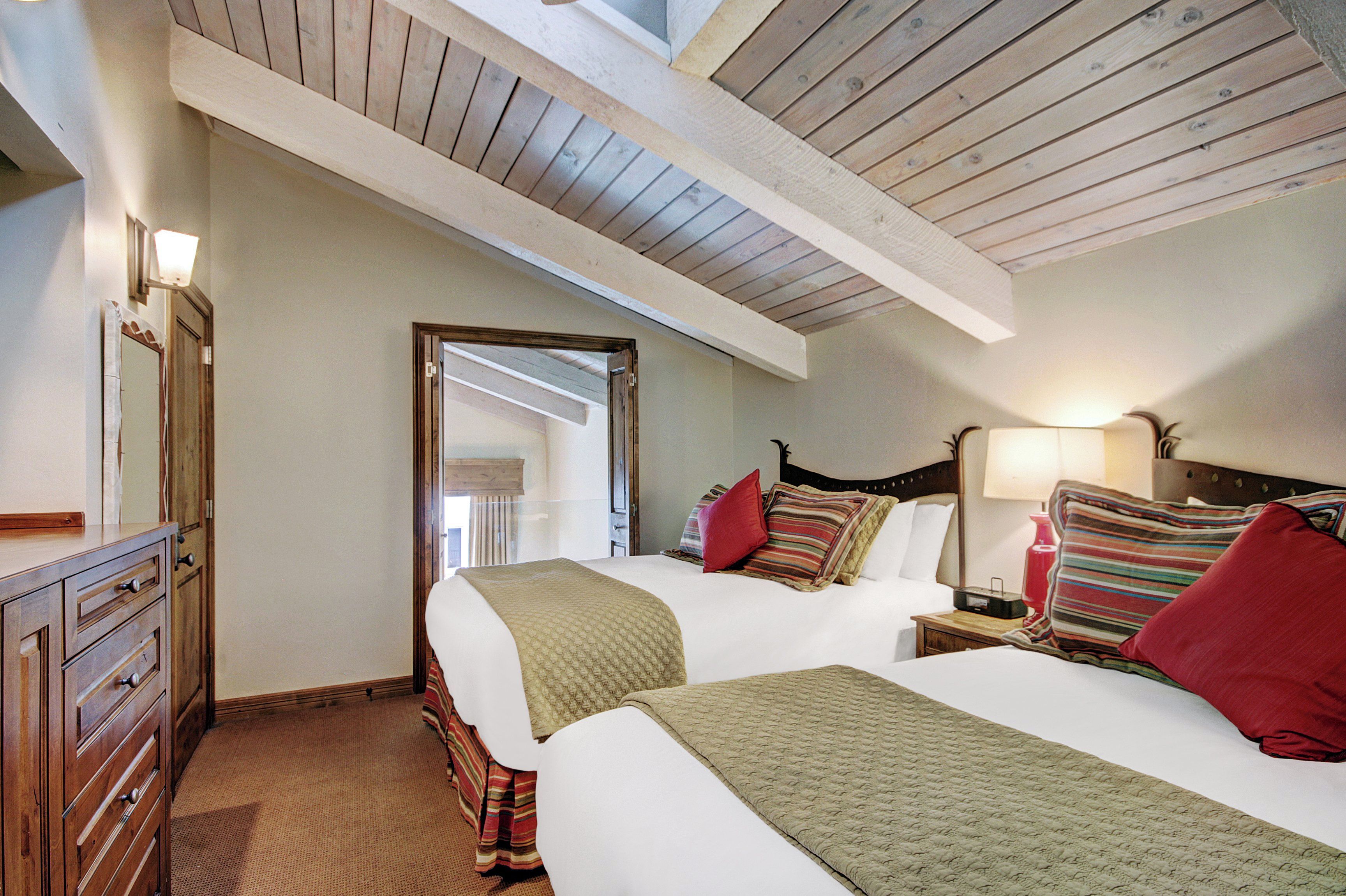 Une chambre avec deux lits, des poutres apparentes en bois au plafond, une commode et une table de chevet avec une lampe.
