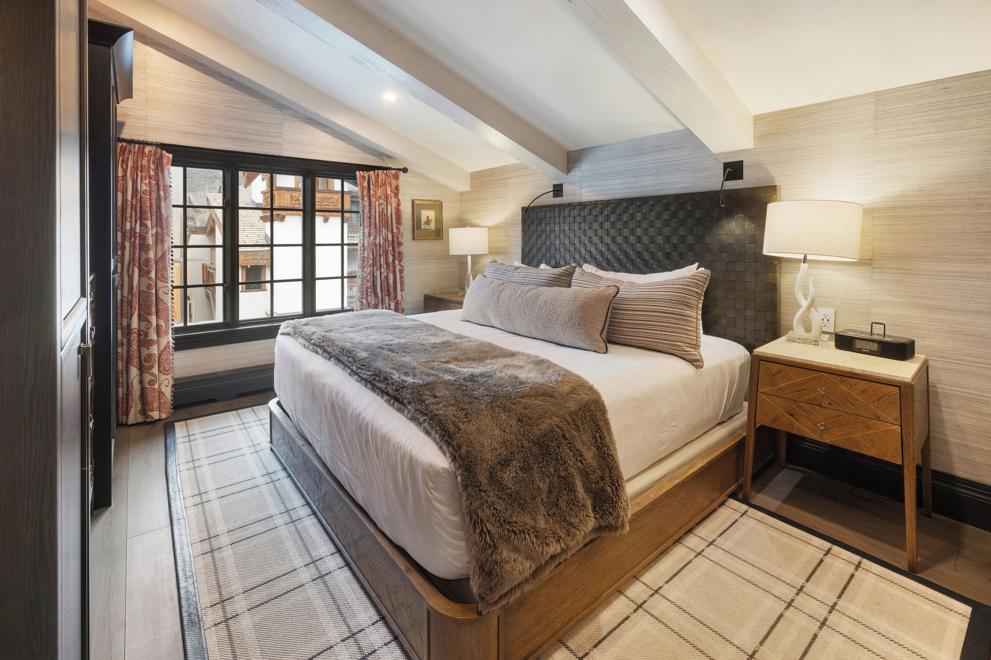 A bedroom with a large bed, a nightstand, a window with curtains, and a ceiling with exposed beams.